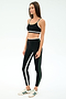 A woman stands in a studio wearing SPLITS59’s Sam High Waist Rigor 7/8 leggings in Black/Military, paired with a matching black run tight sports bra and white sneakers featuring green stripes.