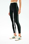 A person wears SPLITS59 Sam High Waist Rigor 7/8 leggings in Black/Military, featuring white and green side stripes, paired with white Adidas sneakers with green stripes, standing against a plain background.