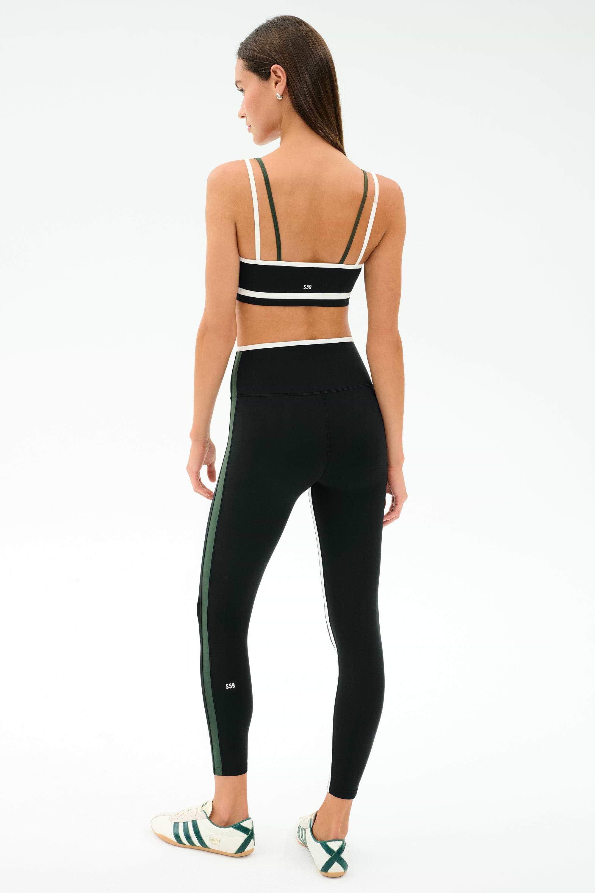 A woman stands with her back to the camera, wearing the SPLITS59 Eva Rigor Double Strap Bra in Black/Military, matching black leggings with side stripes, and white sneakers featuring green accents.