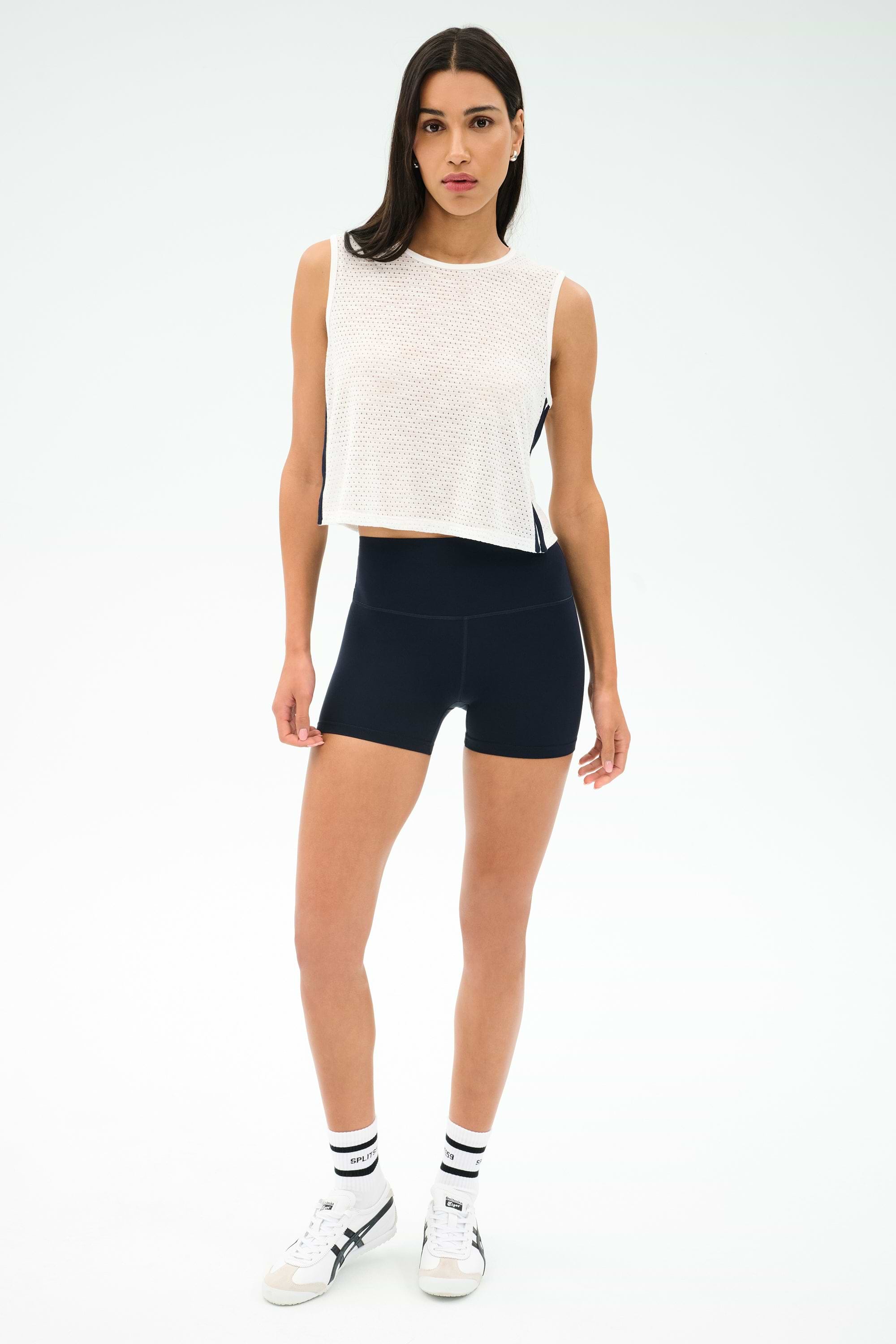 A woman stands against a white background wearing the SPLITS59 Logan Mesh Tank With Stripe in White/Indigo, black fitted shorts, white socks, and white sneakers—an ideal look for breathable sportswear.
