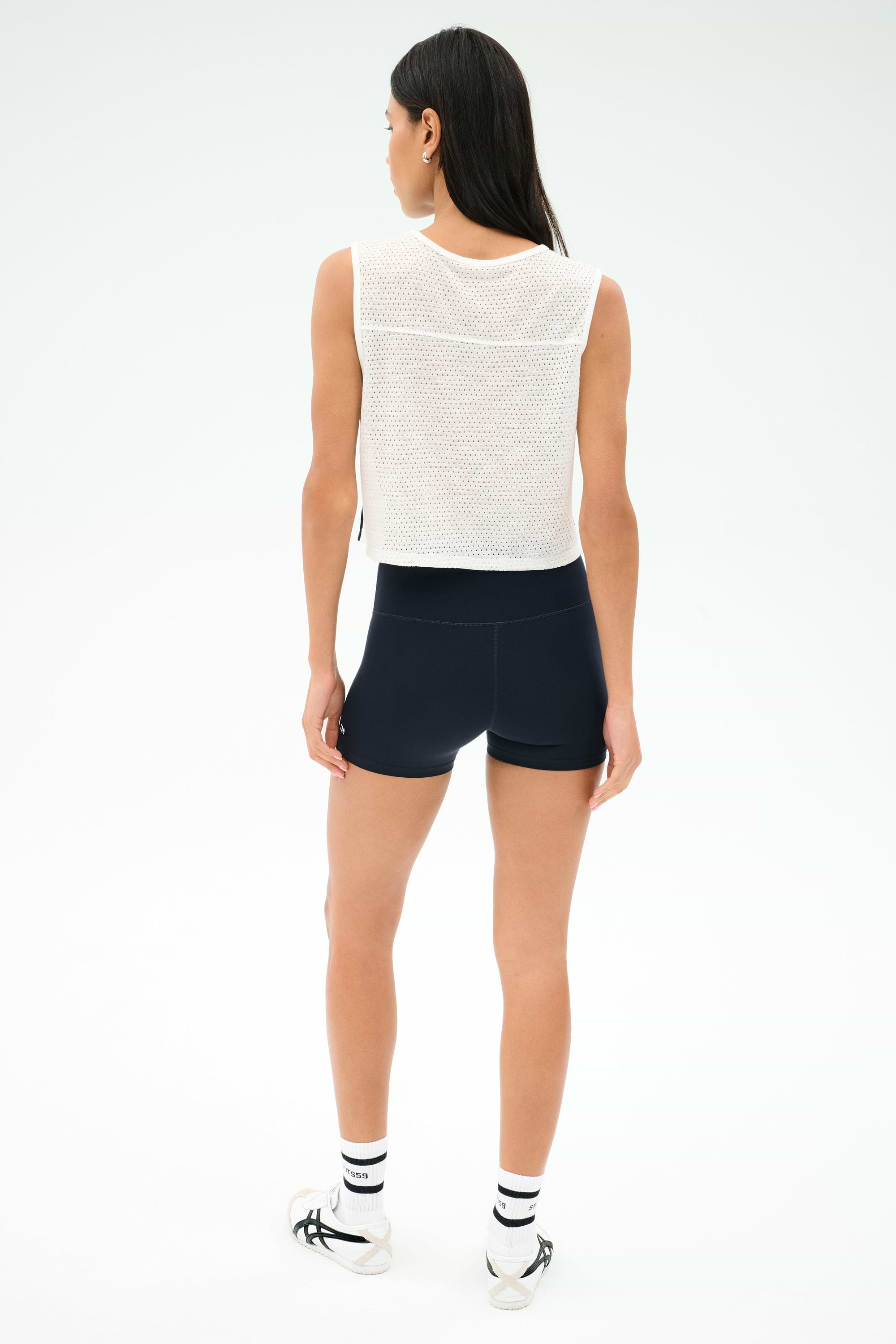 A woman faces away, wearing the SPLITS59 Logan Mesh Tank With Stripe in White/Indigo as a sleeveless workout top, paired with black shorts, white socks with black stripes, and white athletic shoes against a plain light background.
