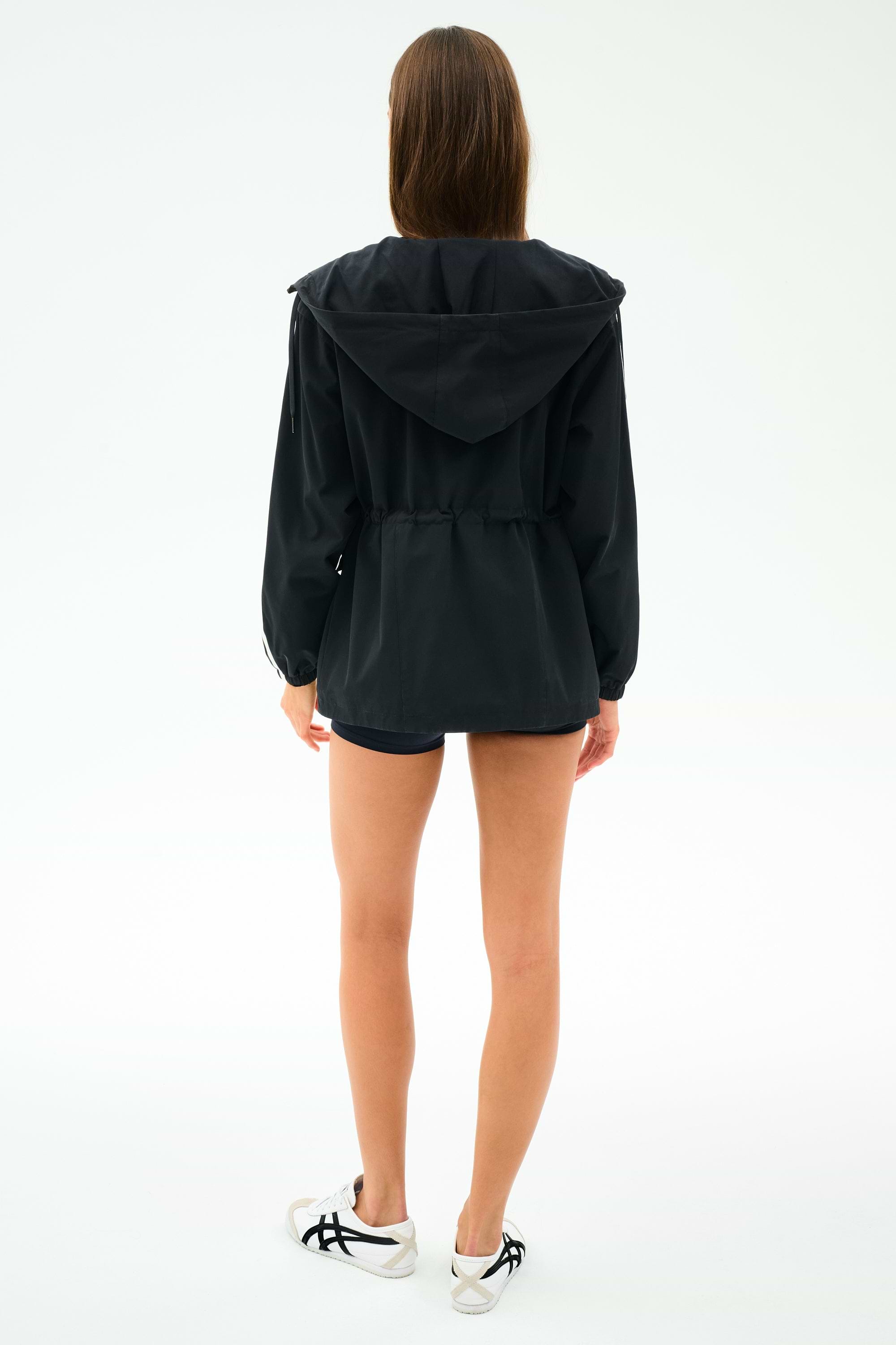 A woman faces away, wearing the SPLITS59 Reggie Water Repellent Jacket in black/white, along with black spandex shorts and white sneakers, against a plain white background.