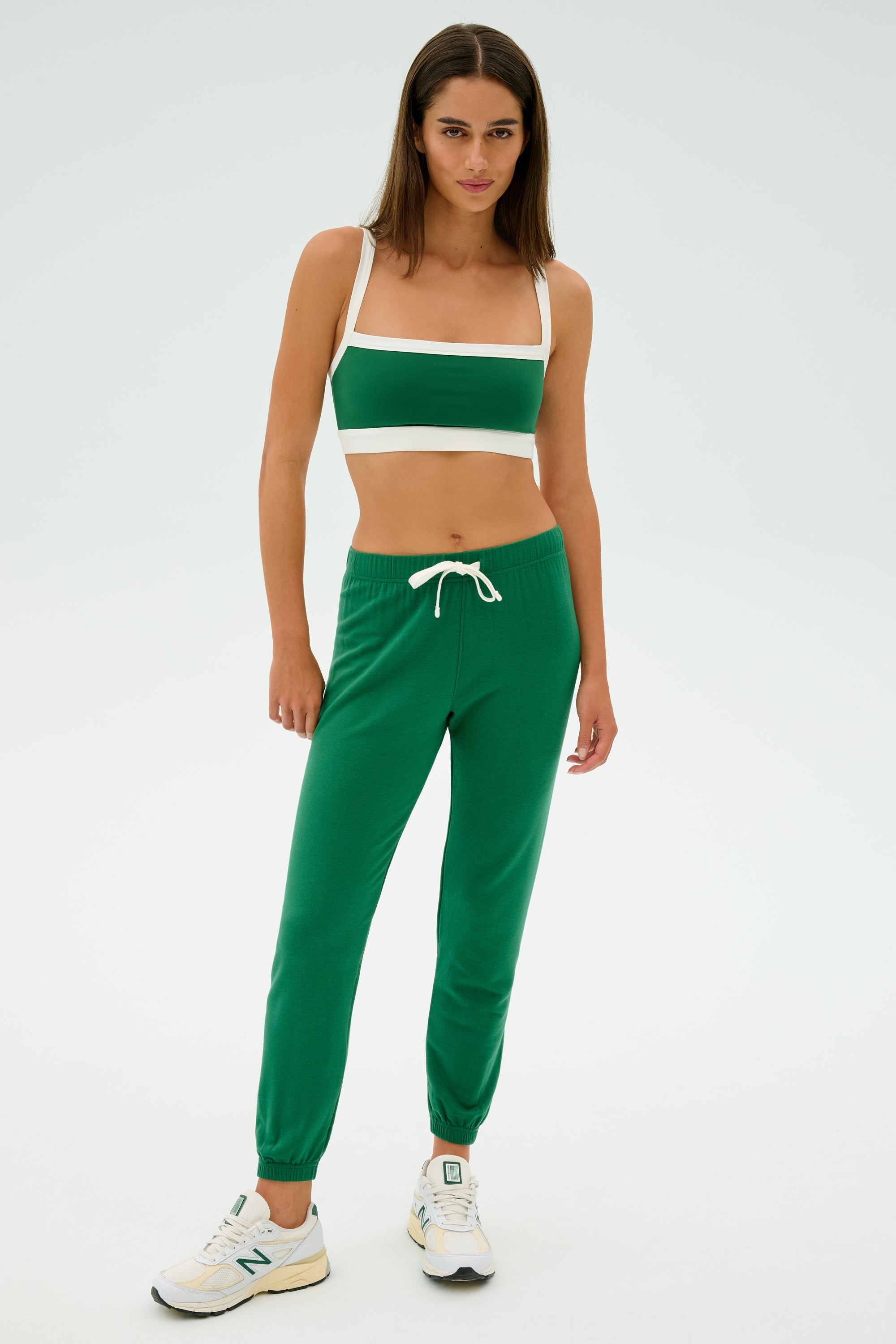 A person stands wearing a green SPLITS59 Sonja Fleece Sweatpant paired with a matching sports bra and white sneakers, against a plain background.