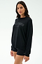 A woman wears the SPLITS59 Andie Oversized Fleece Varsity Graphic Sweatshirt in Black/Splits59 Los Angeles and black shorts, made in Los Angeles, standing against a plain light background.