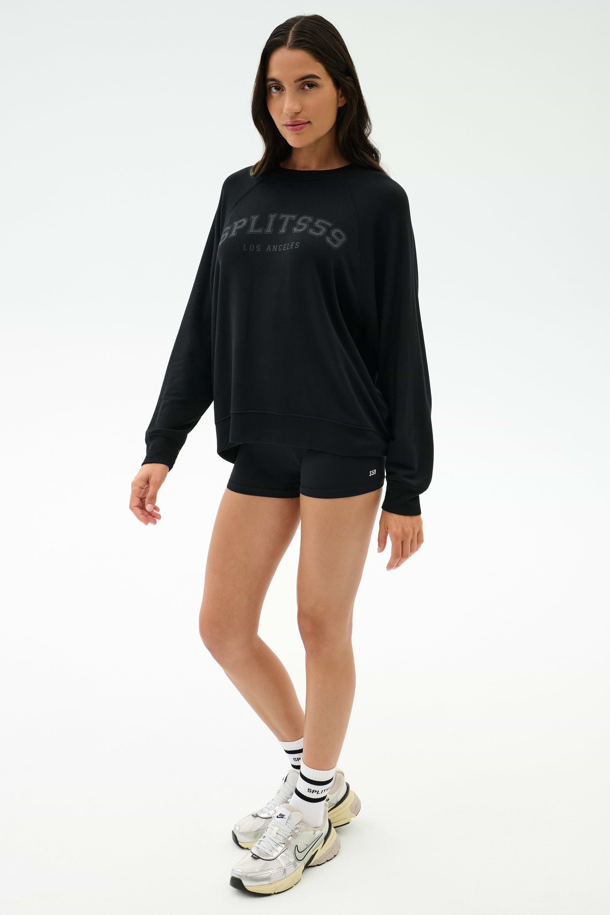 A woman stands in a studio wearing the SPLITS59 Andie Oversized Fleece Varsity Graphic Sweatshirt in black, black shorts, white sneakers, and white socks.
