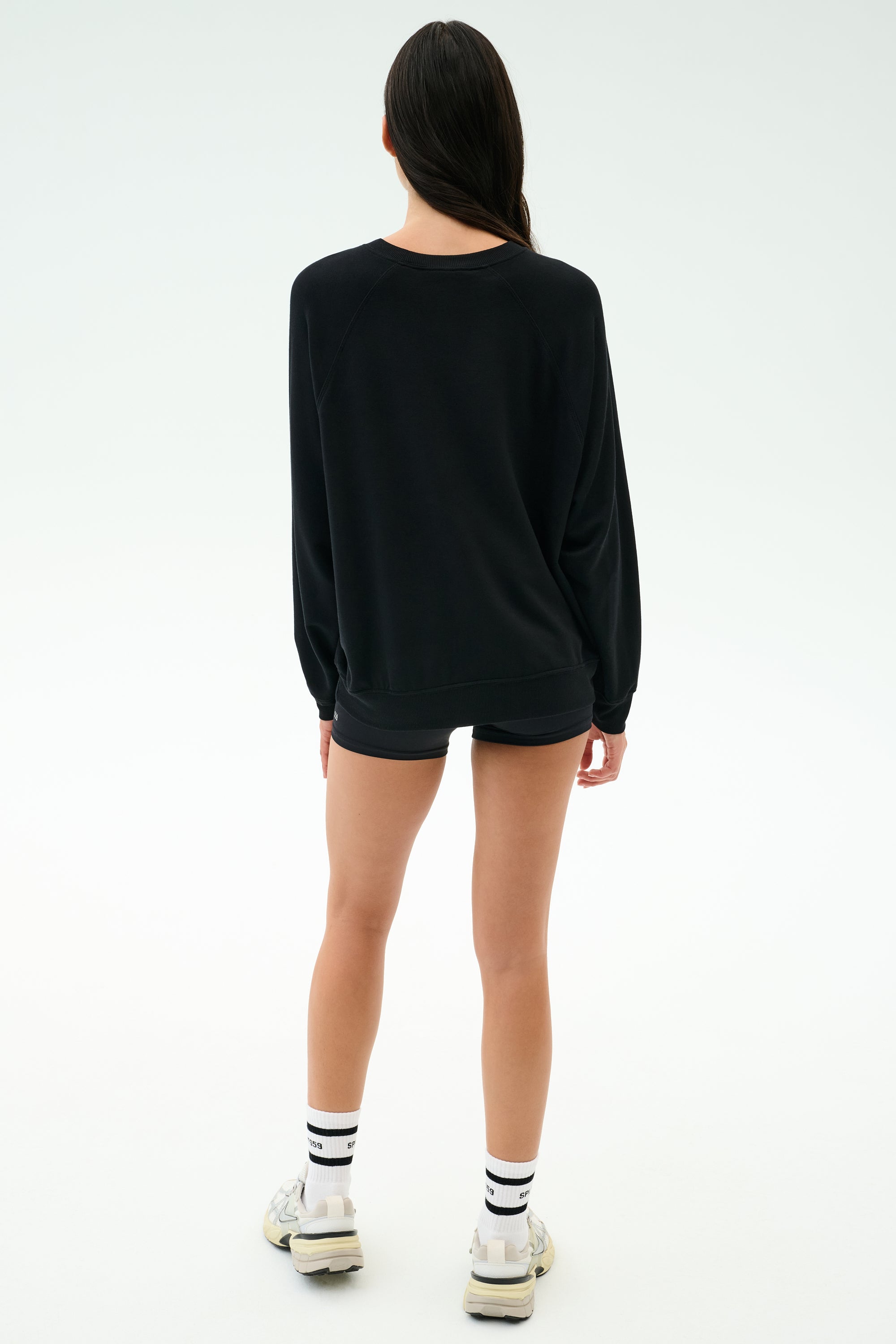 A person faces away, wearing the SPLITS59 Andie Oversized Fleece Varsity Graphic Sweatshirt in black with shorts, white socks with black stripes, and white sneakers, set against a plain light background.
