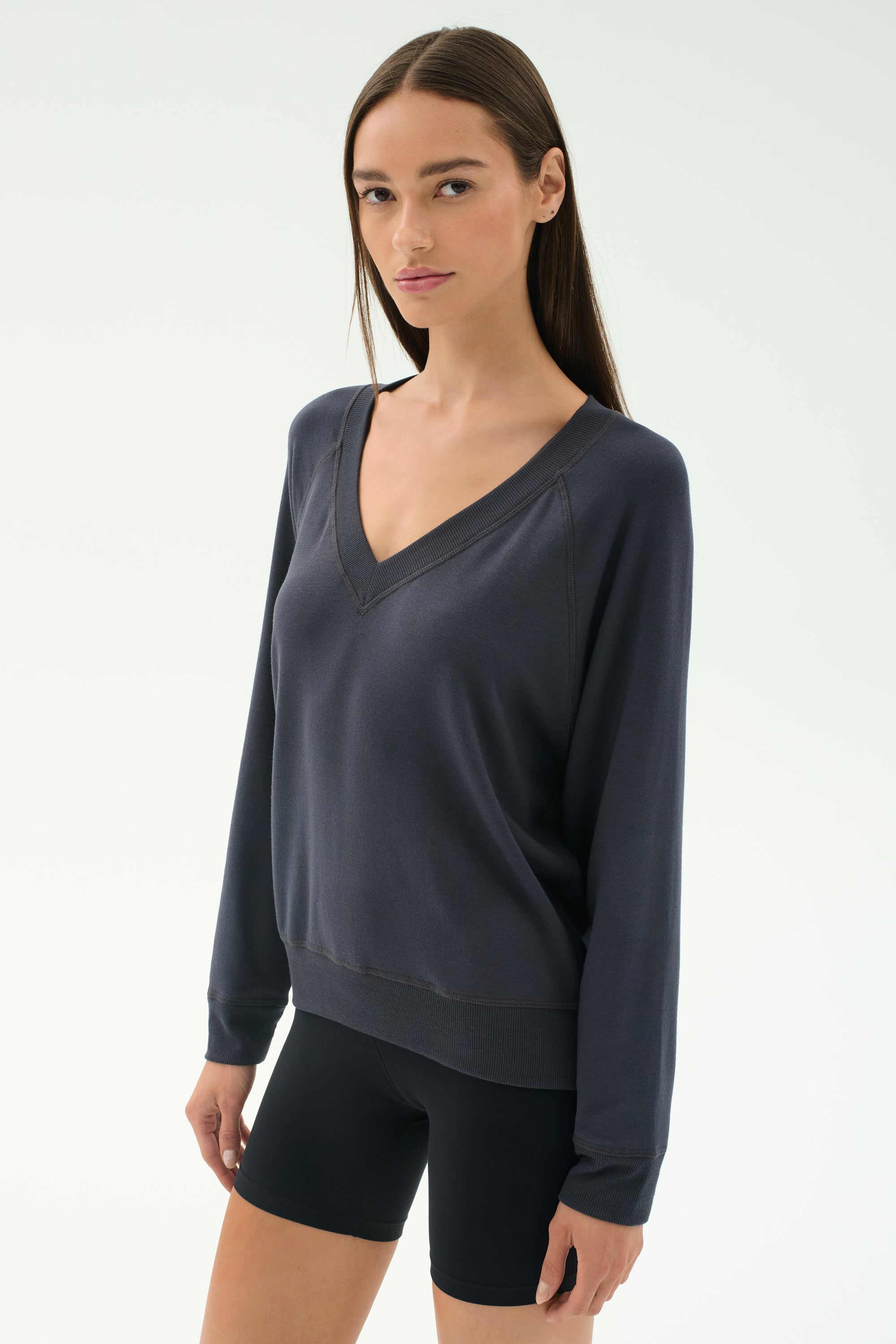A woman with straight brown hair wears the SPLITS59 Bennie Fleece V Neck Sweatshirt in Graphite and black shorts, standing against a plain light background.