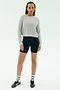 A person with braided hair wears the SPLITS59 Brooks Fleece Long Sleeve Sweatshirt in heather grey and black cycling shorts, paired with striped white socks and black sneakers, standing against a plain white background.
