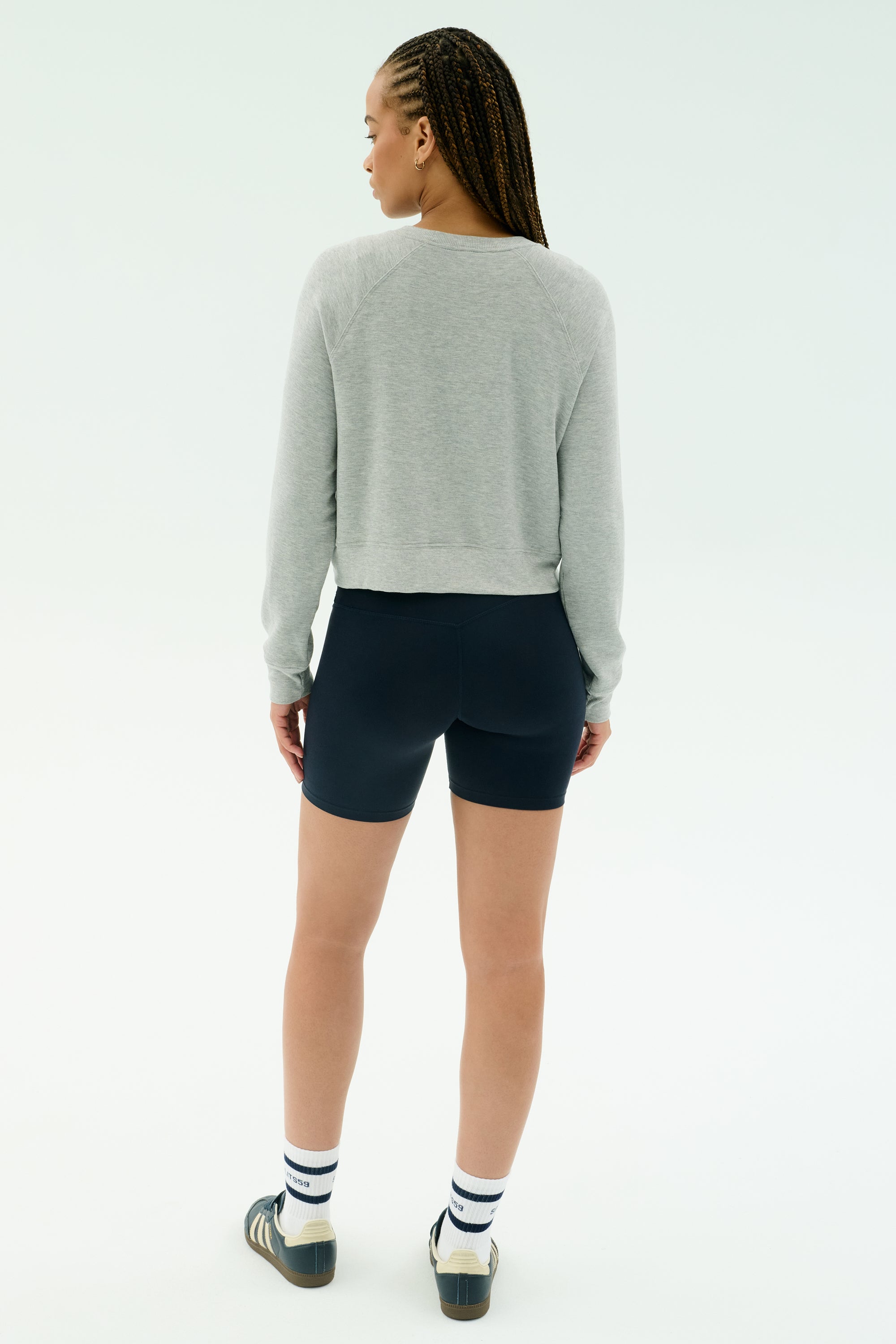 A woman faces away, wearing the SPLITS59 Brooks Fleece Long Sleeve Sweatshirt in Heather Grey, paired with black shorts, striped socks, and sneakers against a plain light background.