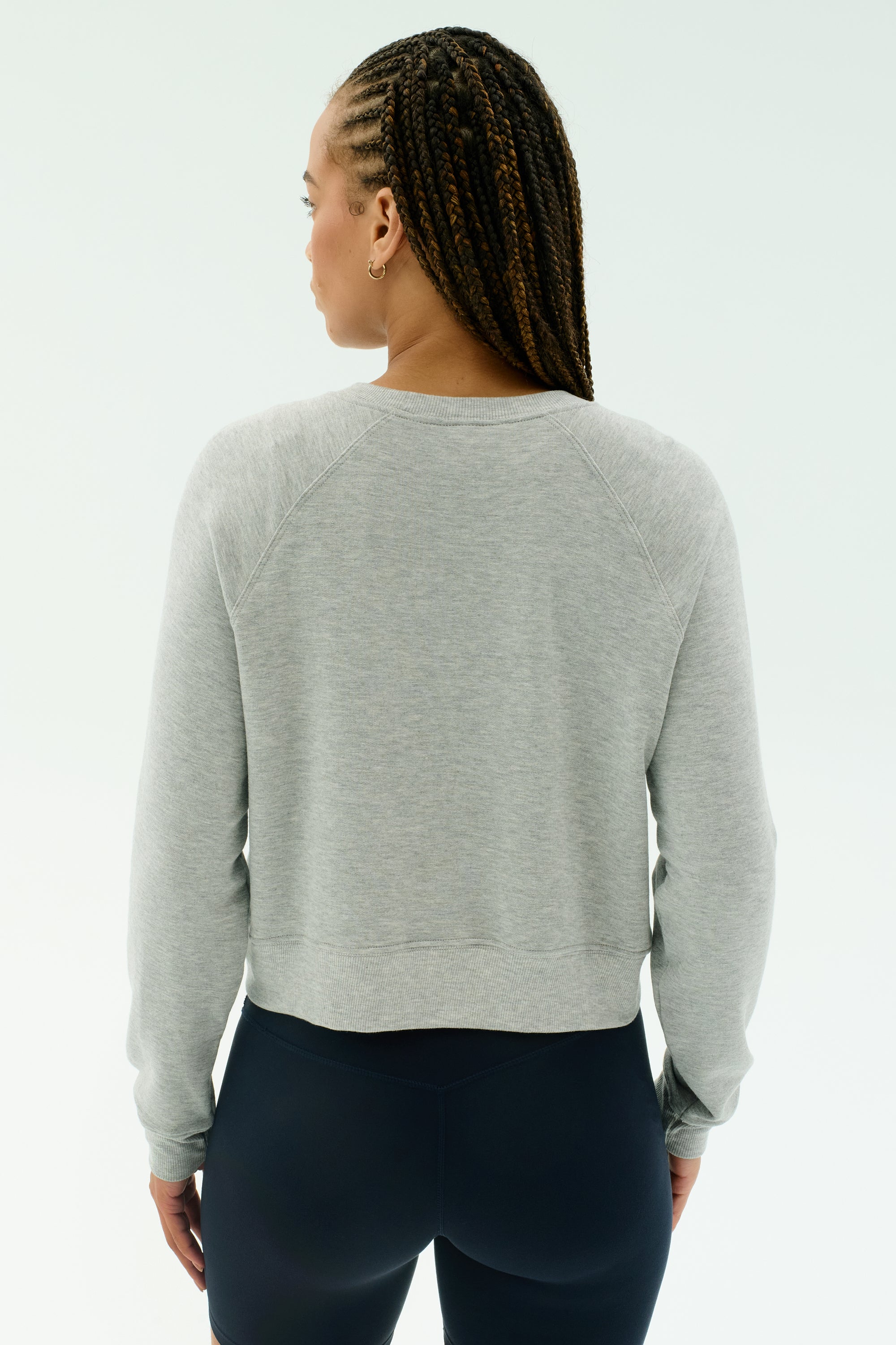 A woman stands with her back to the camera wearing the SPLITS59 Brooks Fleece Long Sleeve Sweatshirt in heather grey, paired with black leggings, set against a plain light background.