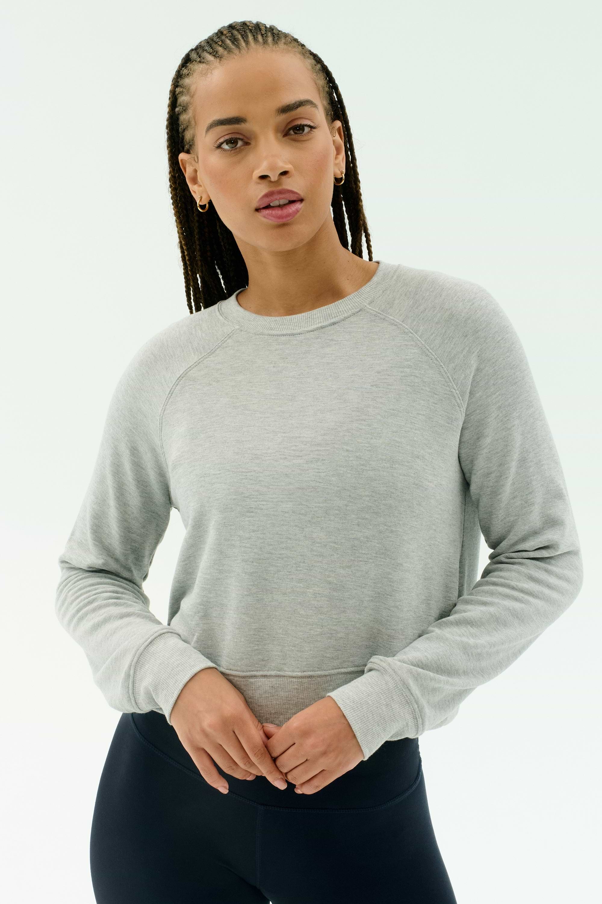 A person with braided hair wears the SPLITS59 Brooks Fleece Long Sleeve Sweatshirt in Heather Grey and black pants, standing against a plain white background.
