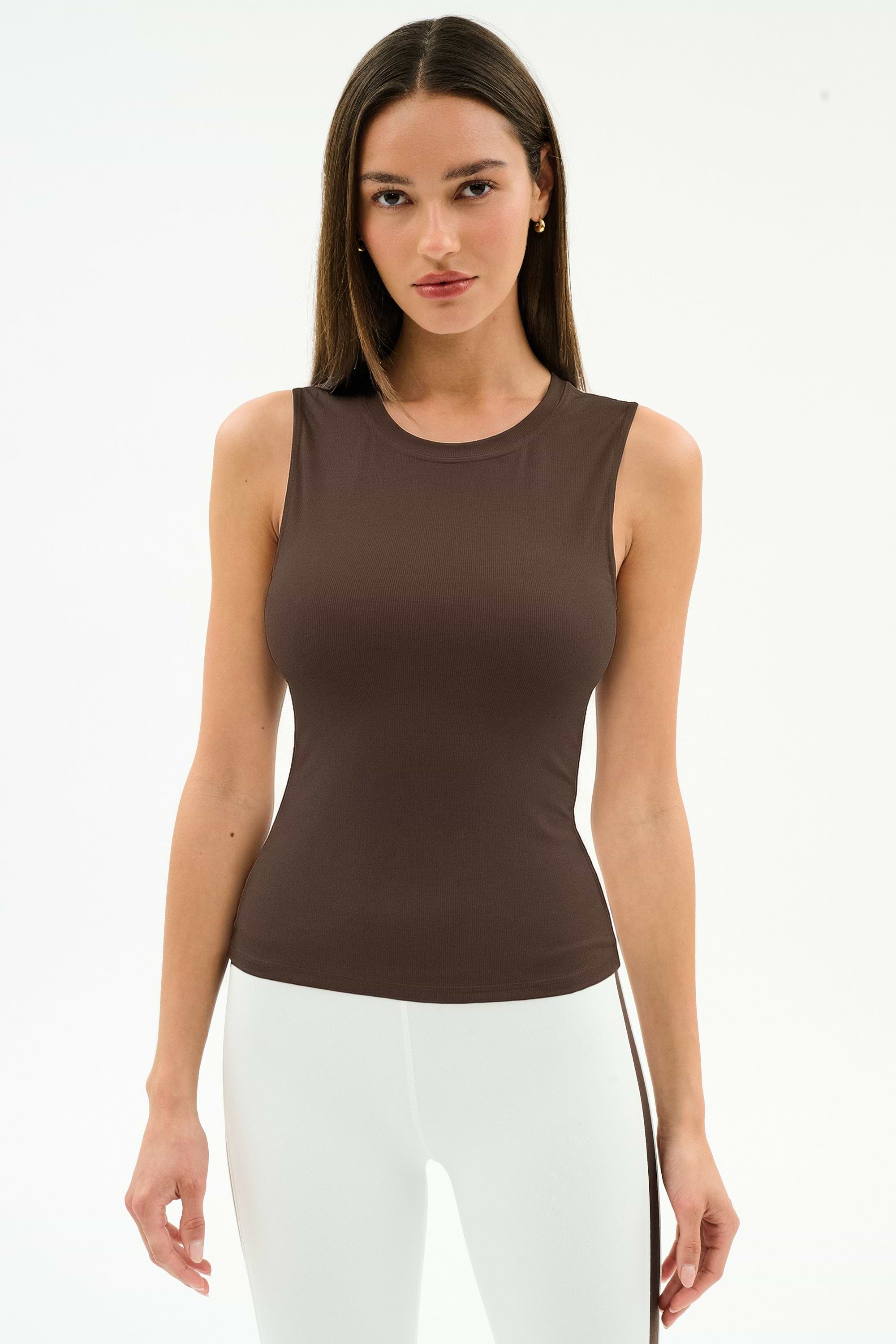 A woman wears the SPLITS59 Carter Airweight Lite Tank in Dark Chocolate with white pants, standing against a plain white background. The buttery soft workout top brings comfort to her minimalist look.