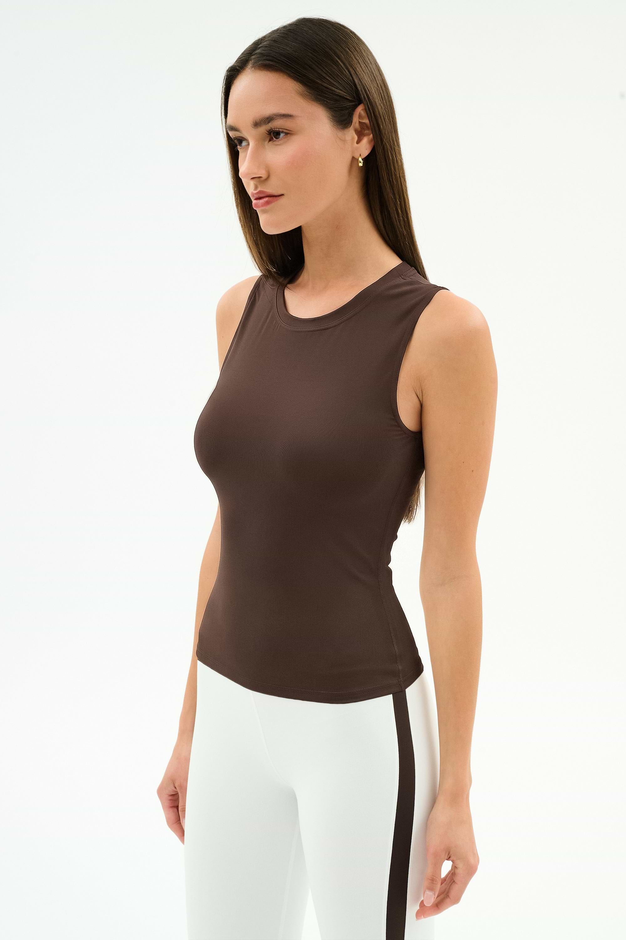 A woman stands against a white background wearing the SPLITS59 Carter Airweight Lite Tank in Dark Chocolate, paired with white pants featuring a dark side stripe.