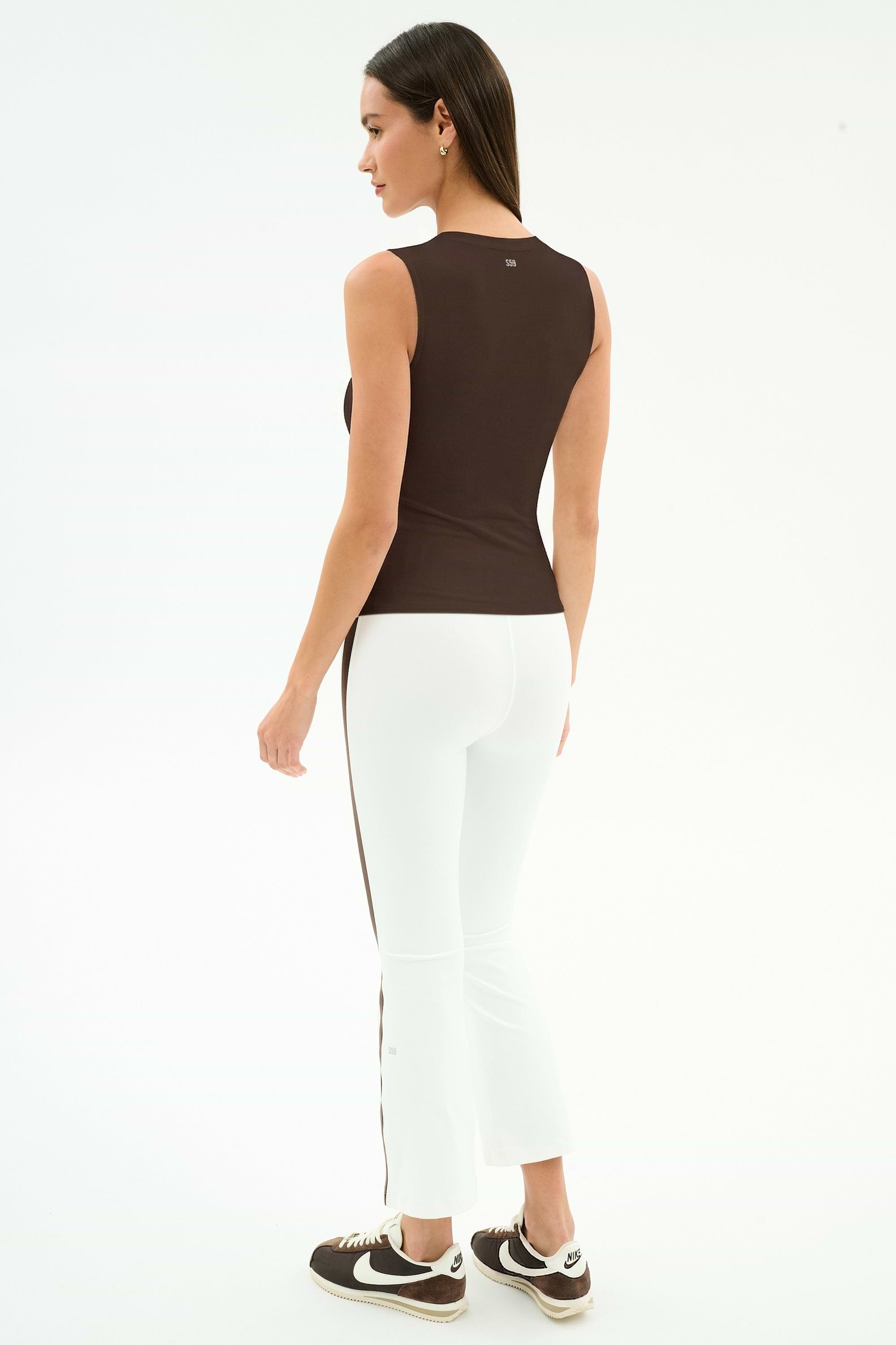 A woman stands facing away, wearing a sleeveless dark brown top, SPLITS59 Raquel High Waist Supplex Crop leggings in White/Dark Chocolate, and black Nike sneakers against a plain white background.