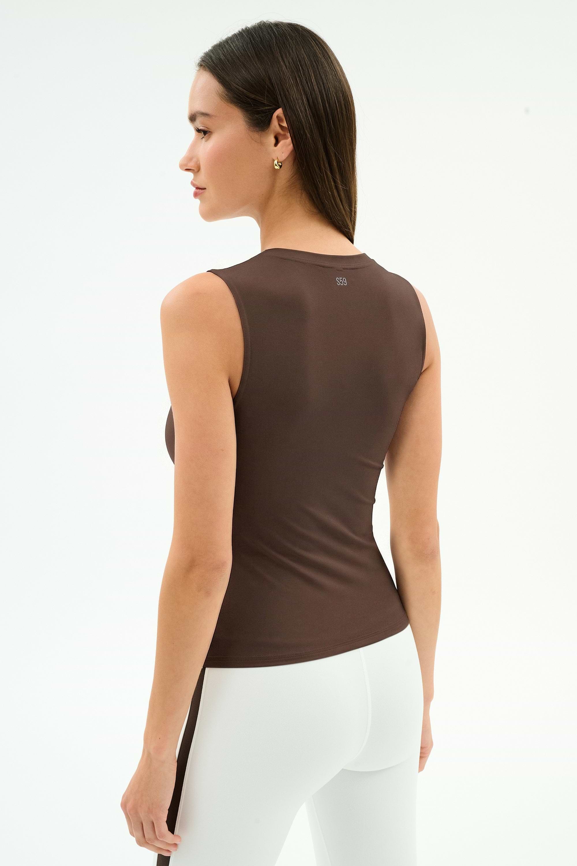 A woman with straight dark hair wears the SPLITS59 Carter Airweight Lite Tank in Dark Chocolate and white pants, facing away in a minimal studio. The tank’s buttery soft fabric adds comfort to her simple look.