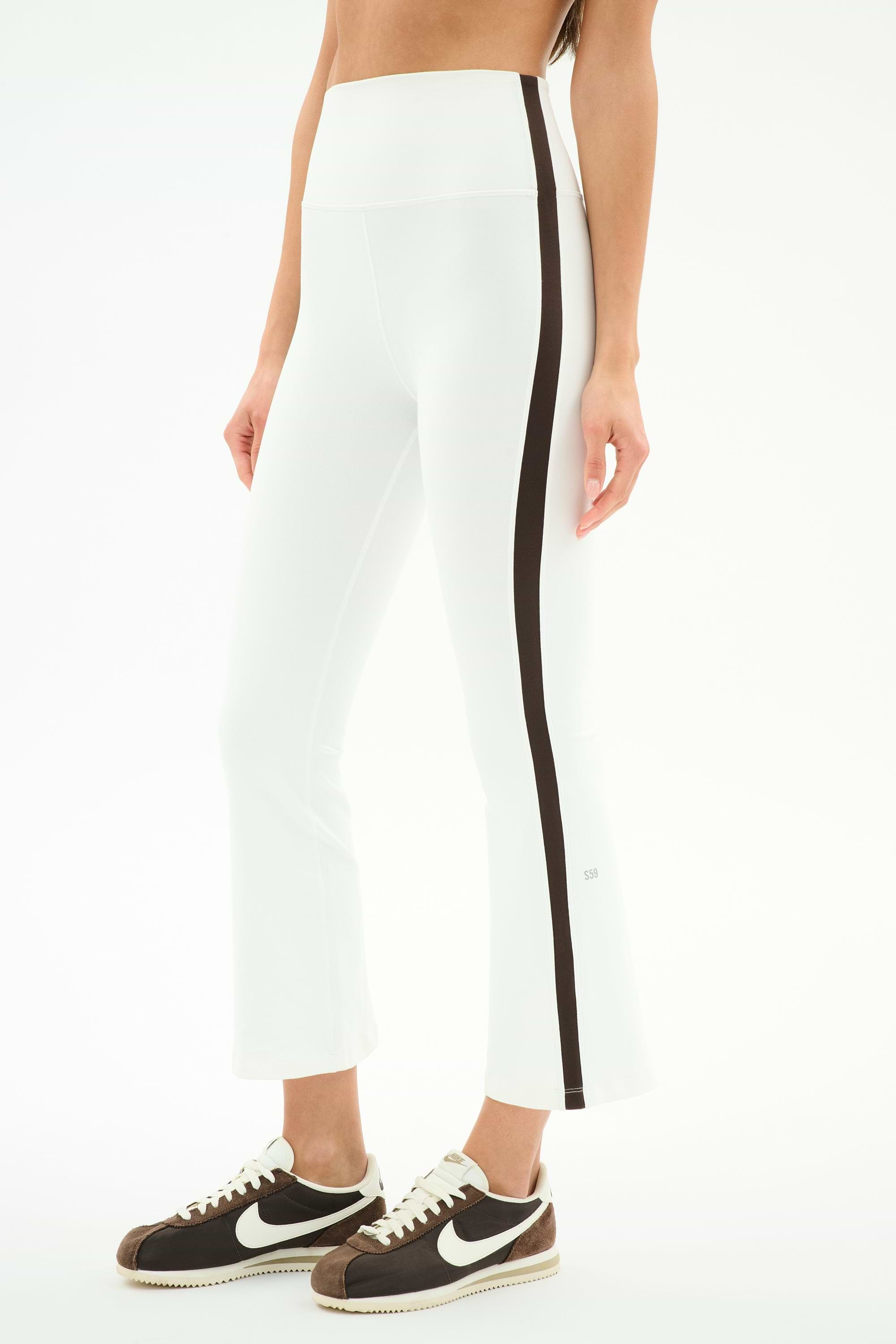 A person wears SPLITS59's Raquel High Waist Supplex Crop in White/Dark Chocolate—high-waisted white leggings with a dark chocolate side stripe—and black-and-white Nike sneakers, standing against a white background.