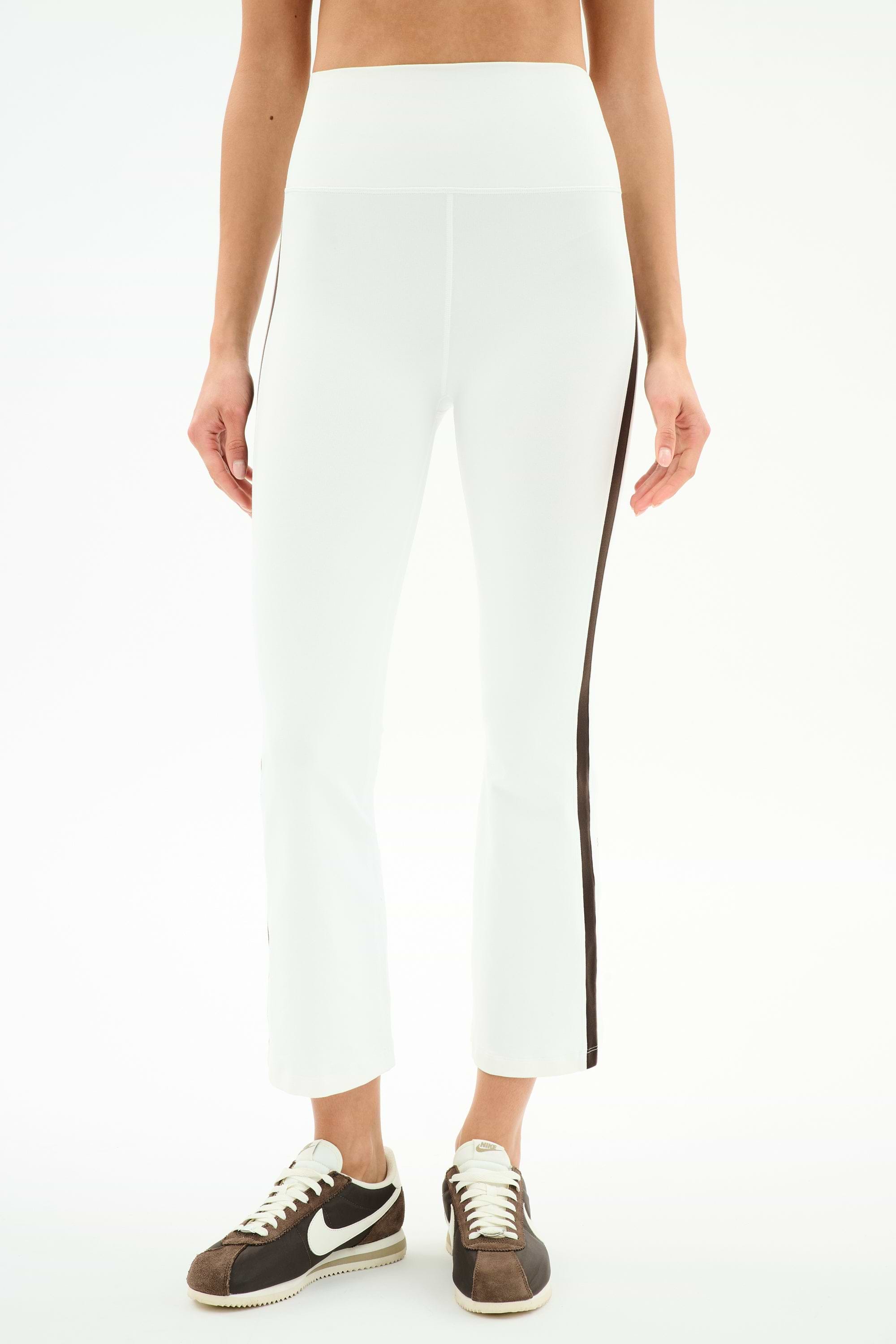 A person wears SPLITS59 Raquel High Waist Supplex Crop pants in White/Dark Chocolate and brown-and-white Nike sneakers, standing against a plain background.