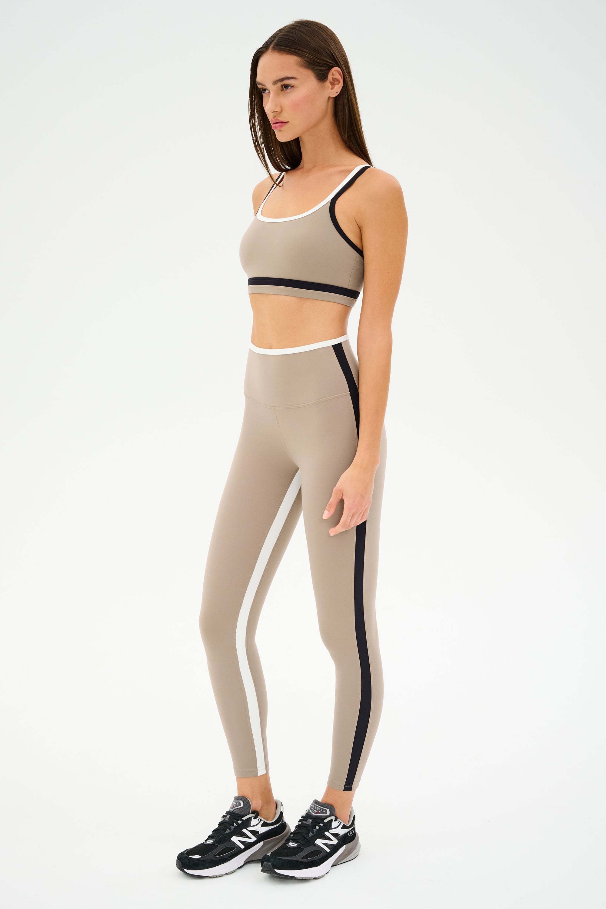 Wearing SPLITS59's Sam High Waist Rigor 7/8 in Latte/Black, one stands poised in a beige athletic ensemble with black accents, exuding workout energy. Black sneakers complete the look as they face slightly right against a simple backdrop.