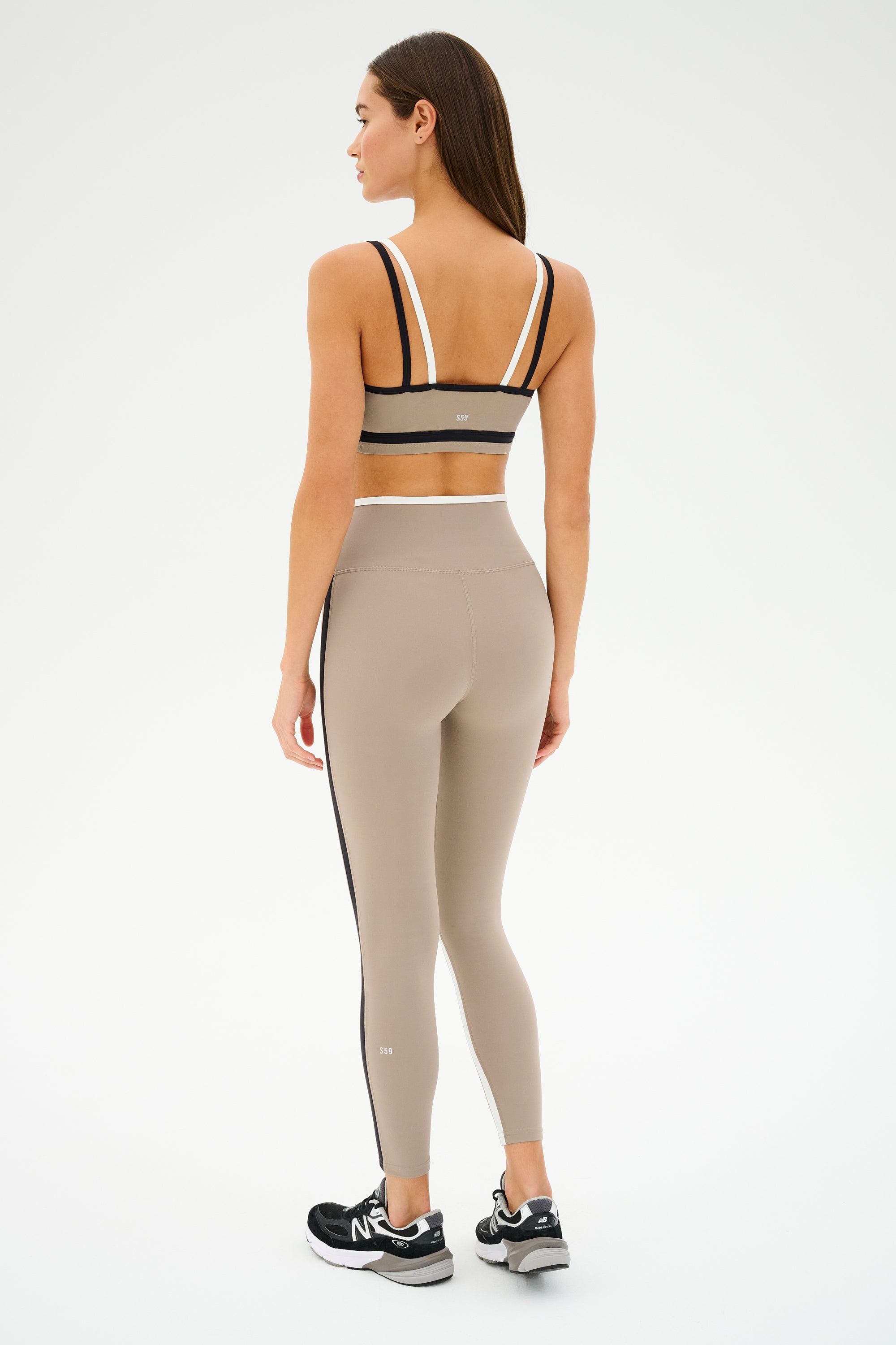 A woman in SPLITS59's Sam High Waist Rigor 7/8 leggings in Latte/Black stands facing away, ready for her yoga session, wearing a black sports bra with white straps and black sneakers with white soles.