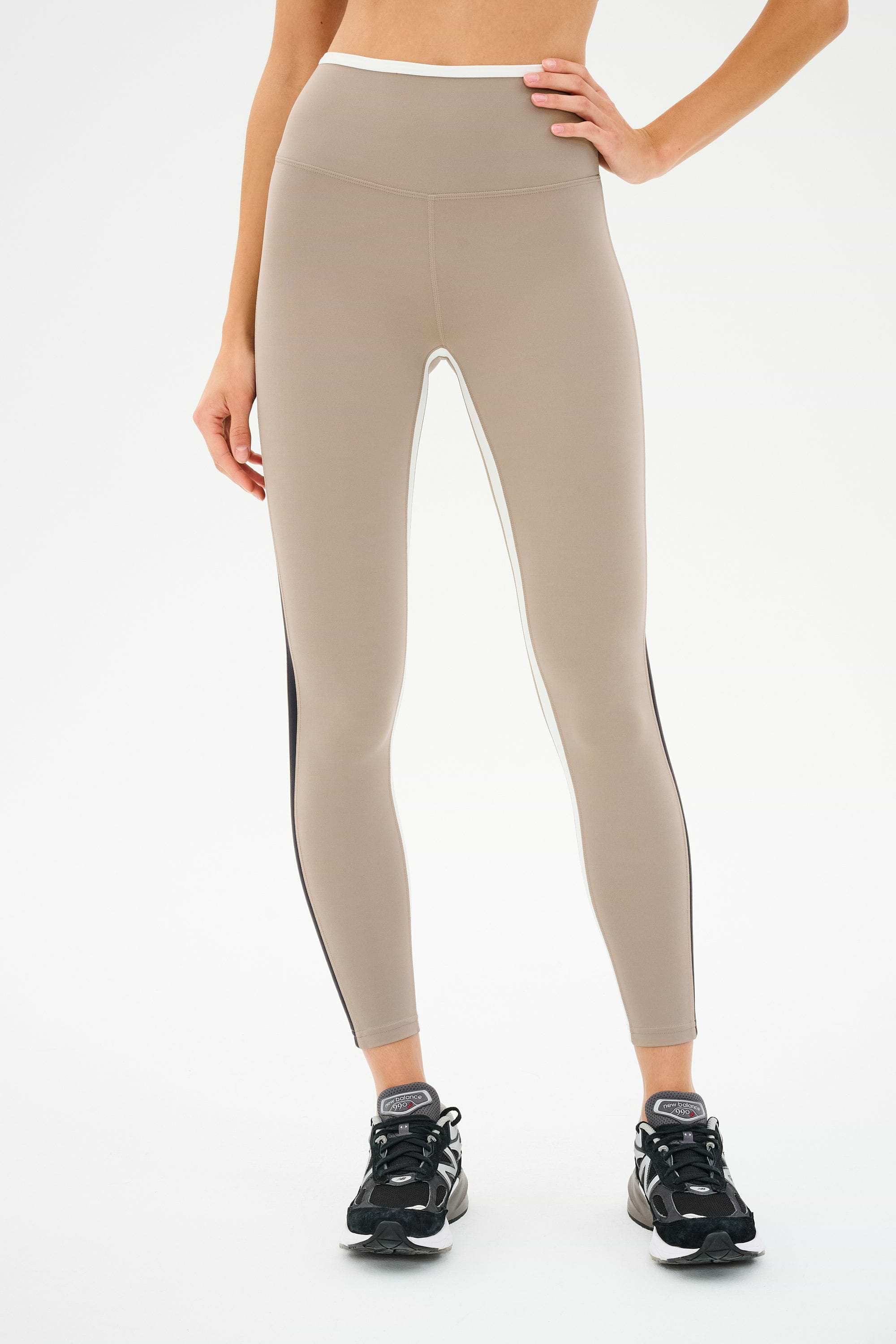 A person wearing SPLITS59 Sam High Waist Rigor 7/8 leggings in Latte/Black with black sneakers, standing against a white background.
