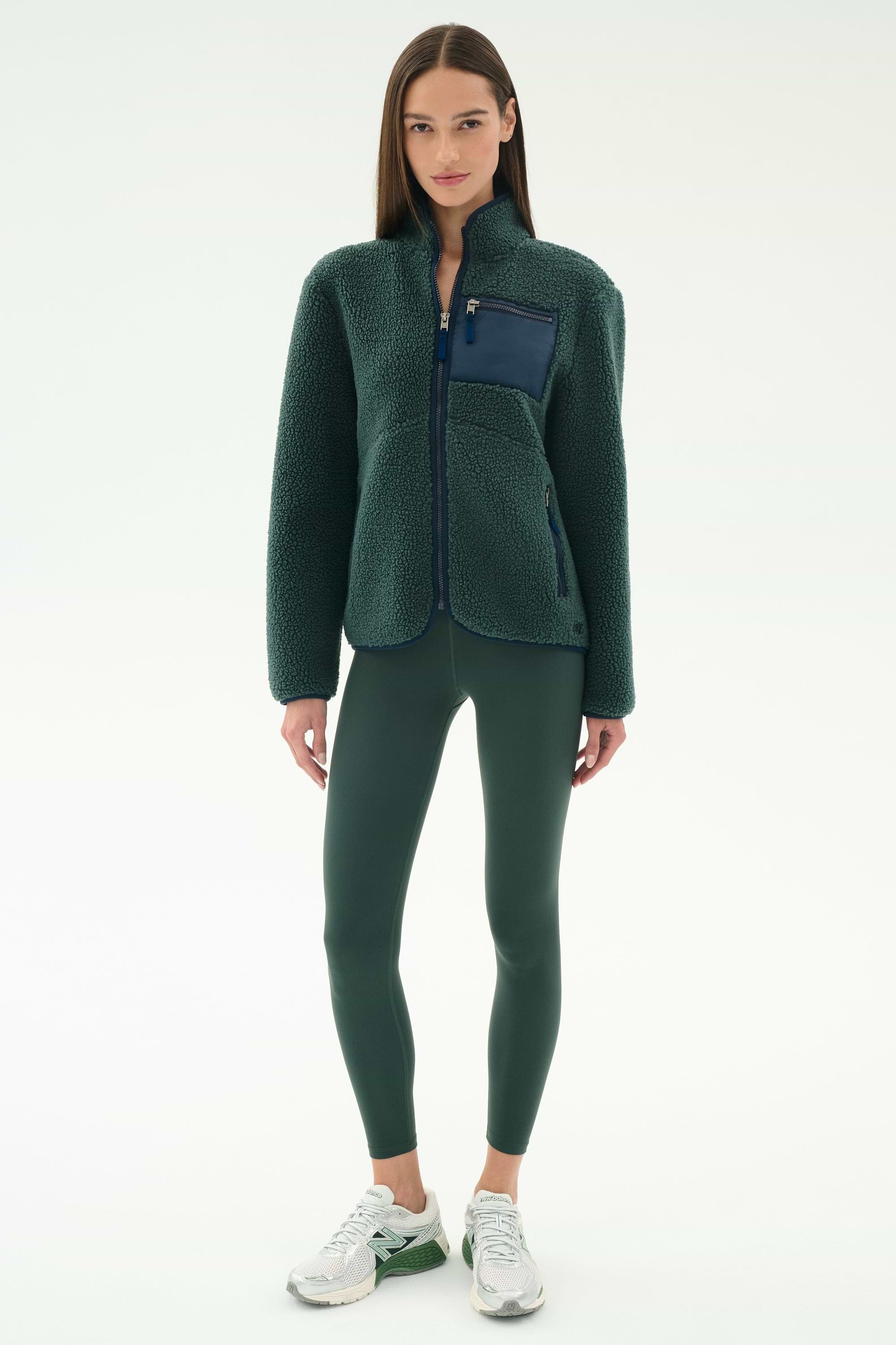 A woman wears the SPLITS59 Harris Sherpa Jacket in Military/Indigo, paired with dark green leggings and white sneakers, standing against a plain light background.