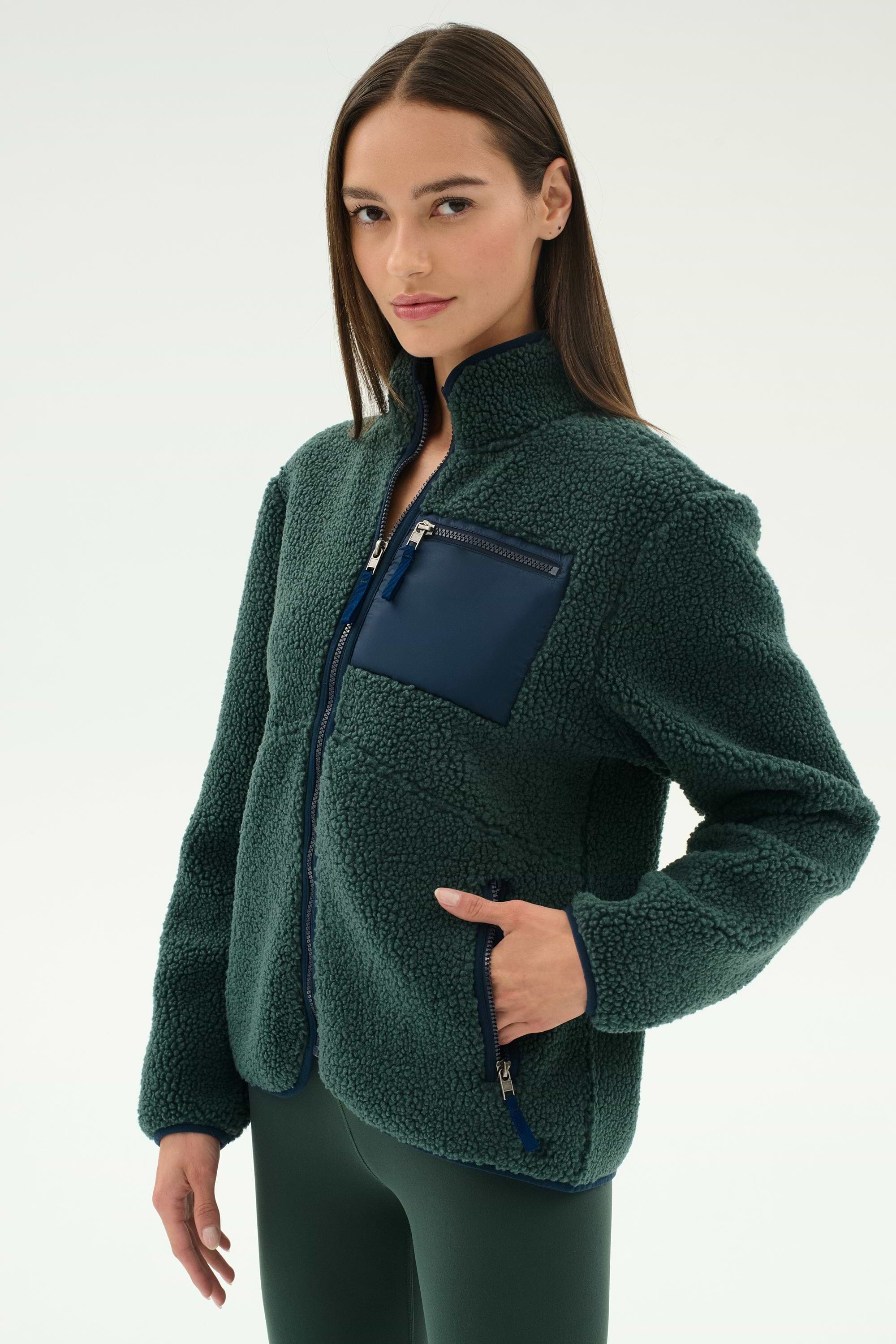 A woman wears the SPLITS59 Harris Sherpa Jacket in Military/Indigo, featuring a zippered navy chest pocket, along with matching pants, standing against a plain light background.