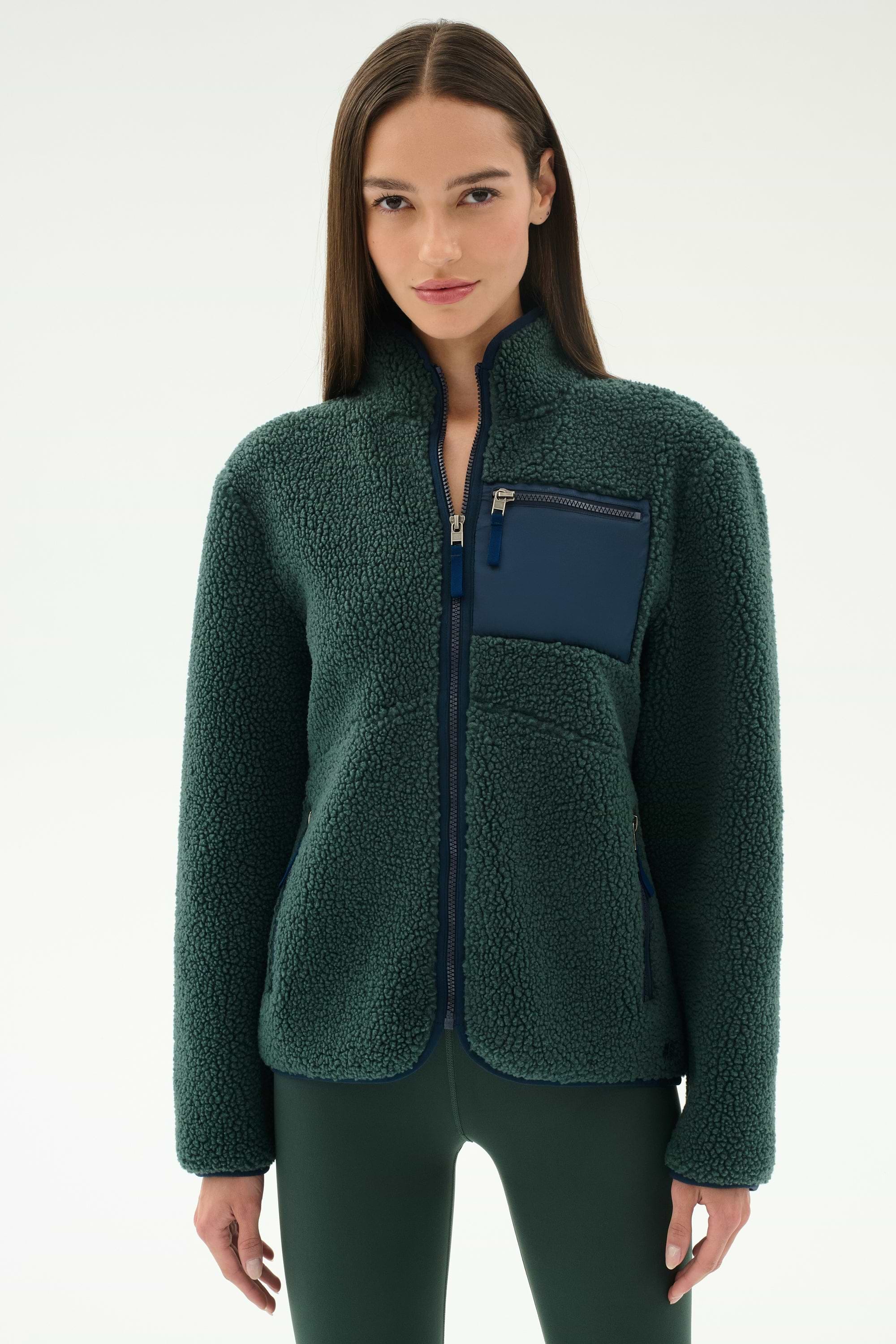 A woman wears the SPLITS59 Harris Sherpa Jacket in Military/Indigo, featuring a navy chest pocket, while standing against a plain light background.