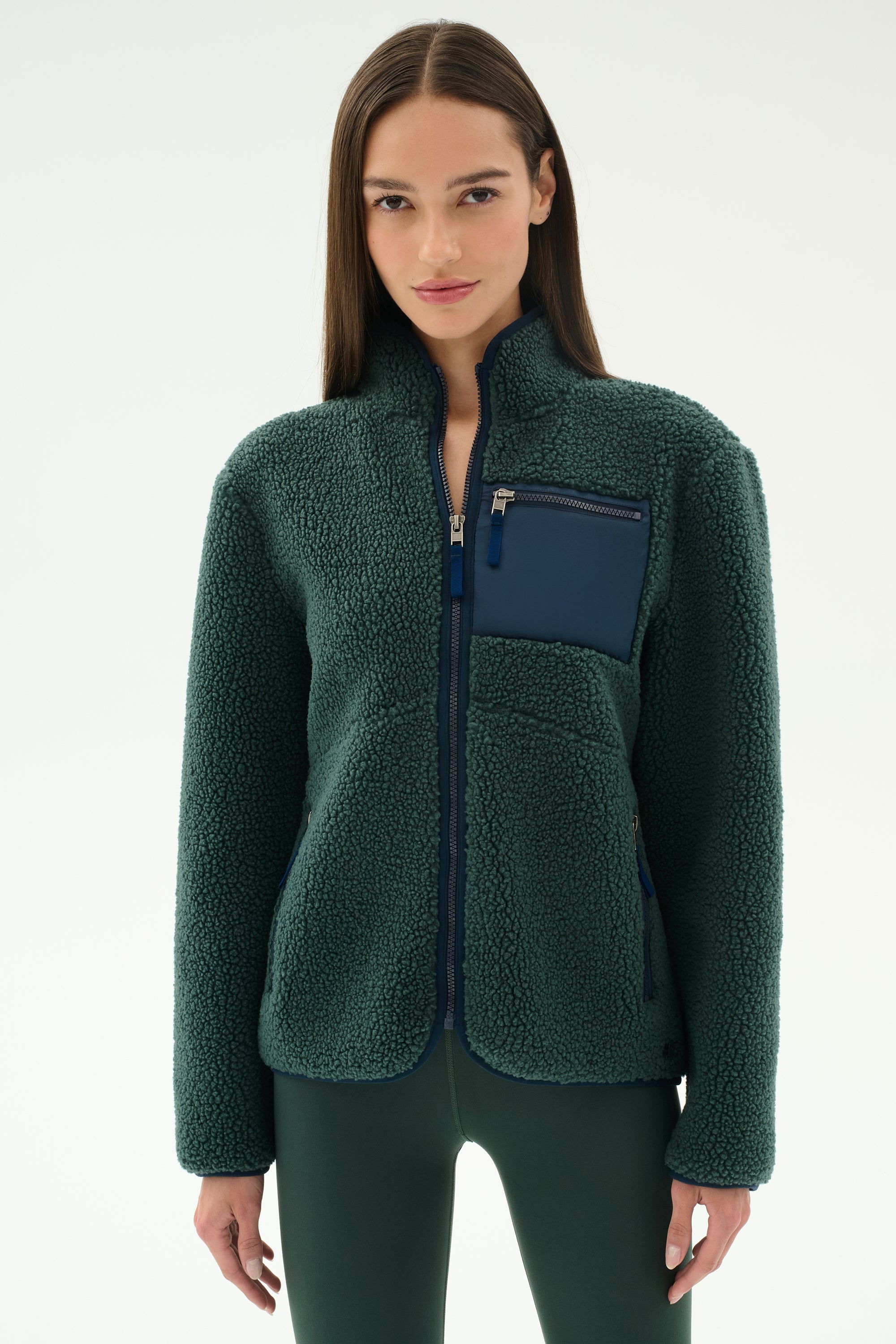 A woman wears the SPLITS59 Harris Sherpa Jacket in Military/Indigo, featuring a navy chest pocket, while standing against a plain light background.