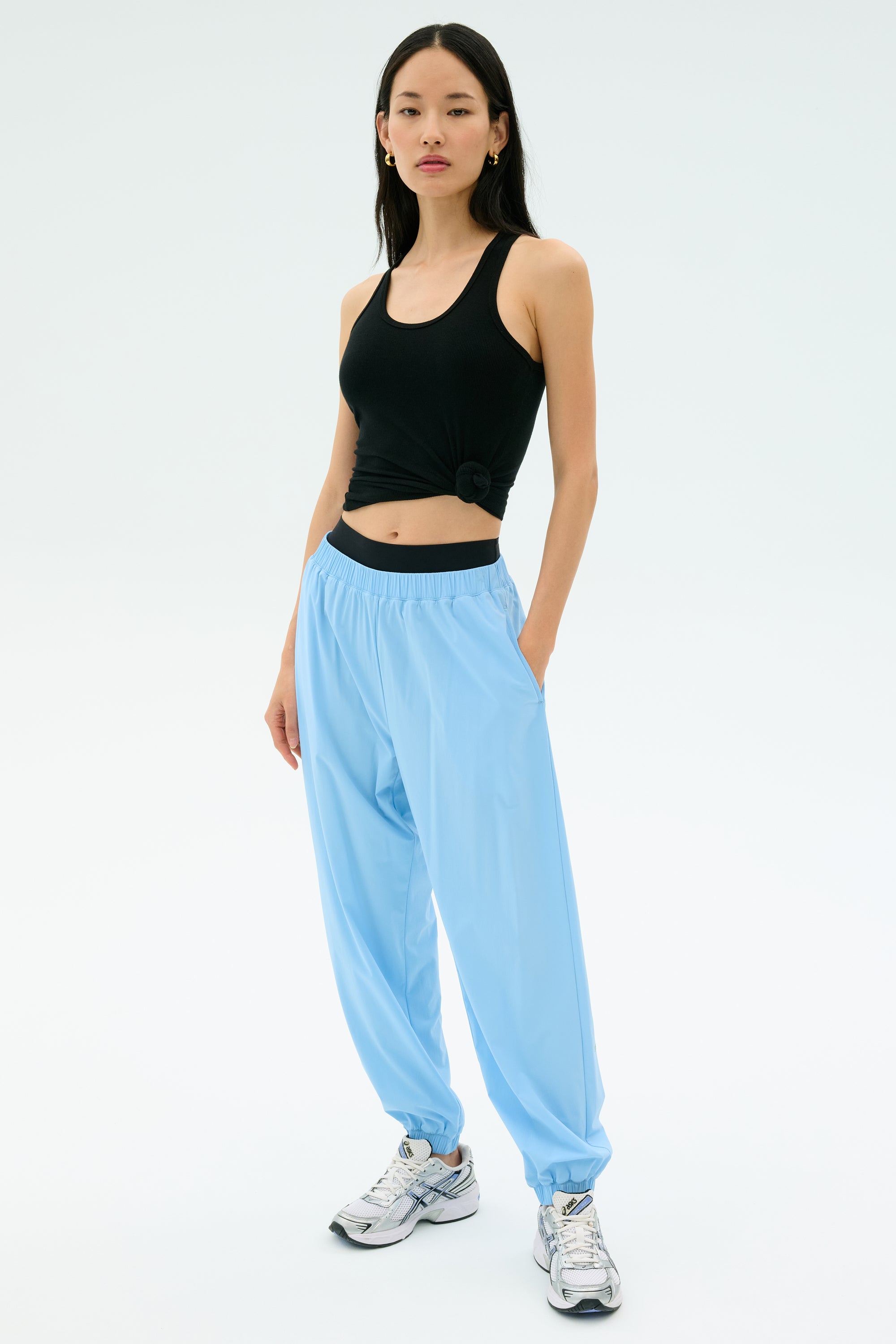 A woman stands against a plain background wearing the Splits59 Ashby Rib Tank in black, paired with light blue jogger pants and white sneakers.