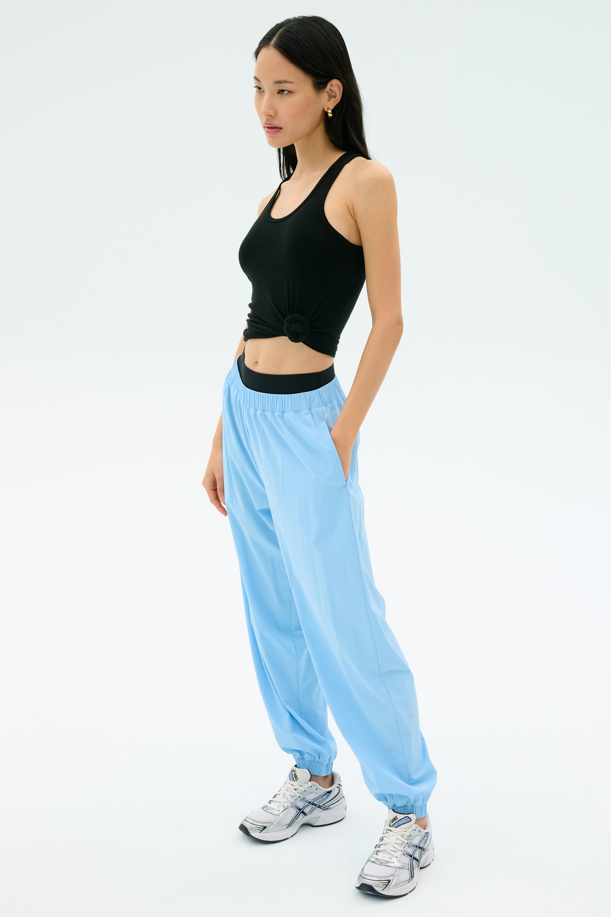 A woman poses against a plain background wearing the Splits59 Ashby Rib Tank in black, paired with light blue joggers and white sneakers—ideal for gym sessions or casual outings.