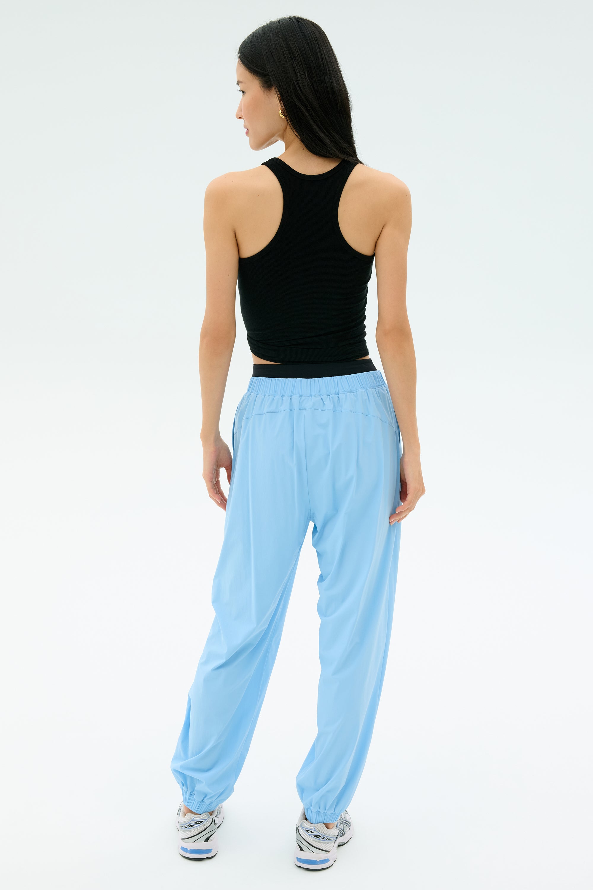 A woman stands facing away, wearing the Splits59 Ashby Rib Tank in black with loose light blue pants and sneakers against a plain light background.