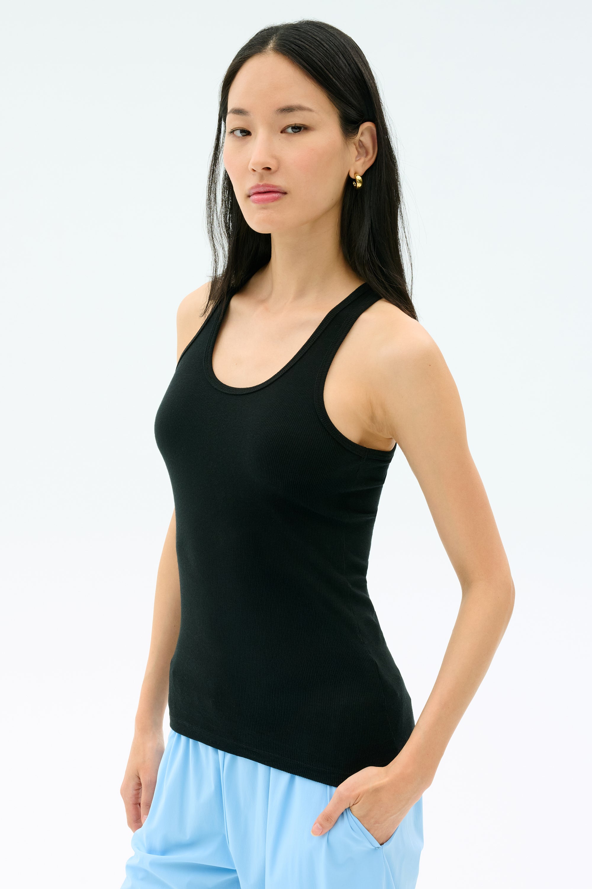 A woman with long dark hair wears the Splits59 Ashby Rib Tank in black and light blue pants, standing against a plain light background.
