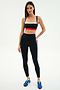A woman stands against a white background wearing a red, white, and blue striped sports bra, SPLITS59 Monah High Waist Rigor Legging 7/8 in Black/Cobalt, and blue sneakers.