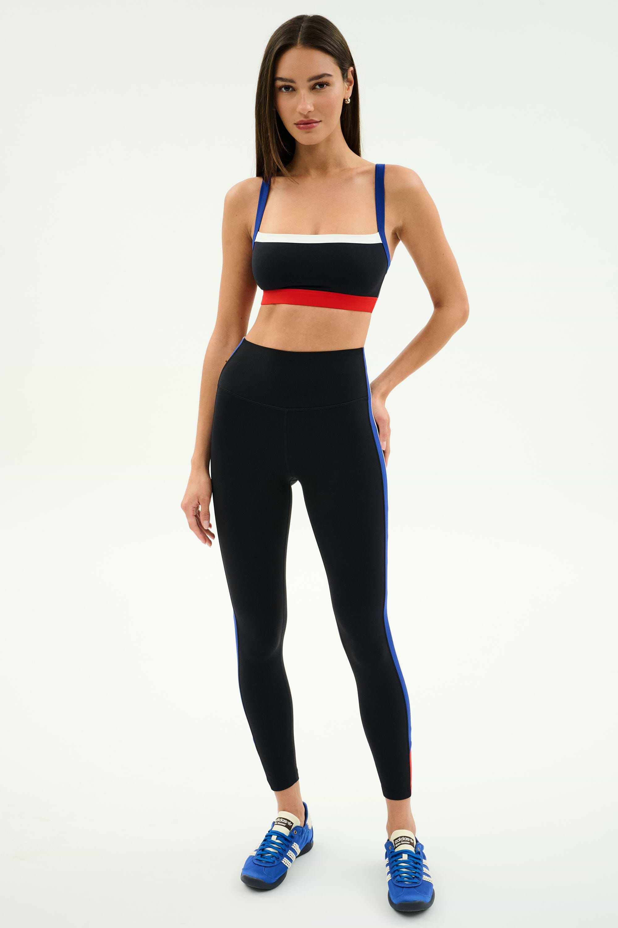A woman stands against a white background wearing the SPLITS59 Monah Rigor Bra in Black/Cobalt, high-waisted black leggings with blue stripes, and blue athletic shoes.