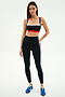 A woman stands against a white background wearing the SPLITS59 Monah Rigor Bra in Black/Cobalt, high-waisted black leggings with blue stripes, and blue athletic shoes.