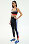 A woman wears SPLITS59's Monah High Waist Rigor Legging 7/8 in Black/Cobalt with blue, white, and red stripes and a matching black sports bra, paired with blue Adidas sneakers against a plain white background.