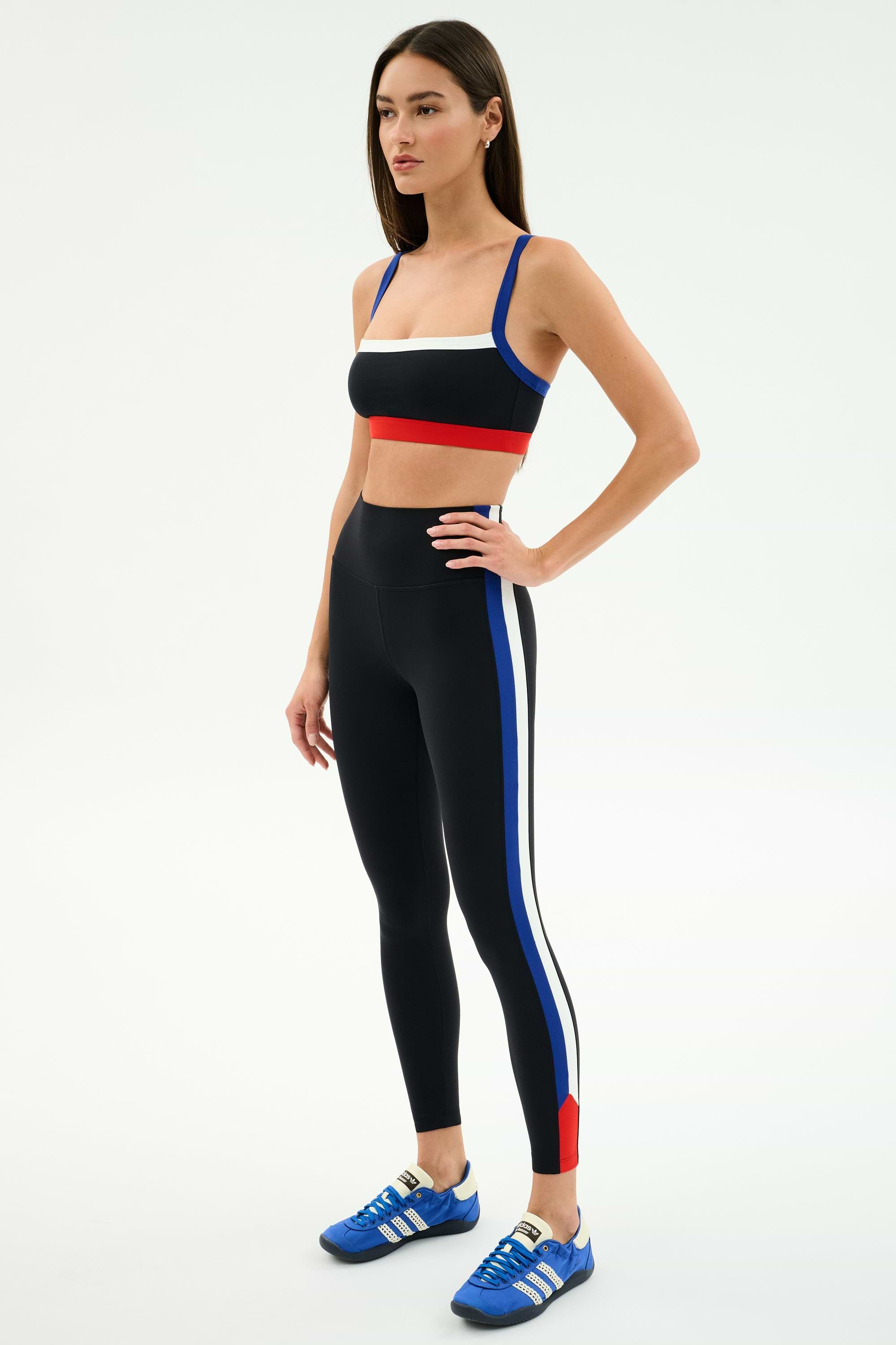 Woman wearing the SPLITS59 Monah Rigor Bra in Black/Cobalt and matching leggings with blue, white, and red stripes, paired with blue athletic shoes, stands against a plain white background.