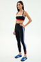 Woman wearing the SPLITS59 Monah Rigor Bra in Black/Cobalt and matching leggings with blue, white, and red stripes, paired with blue athletic shoes, stands against a plain white background.
