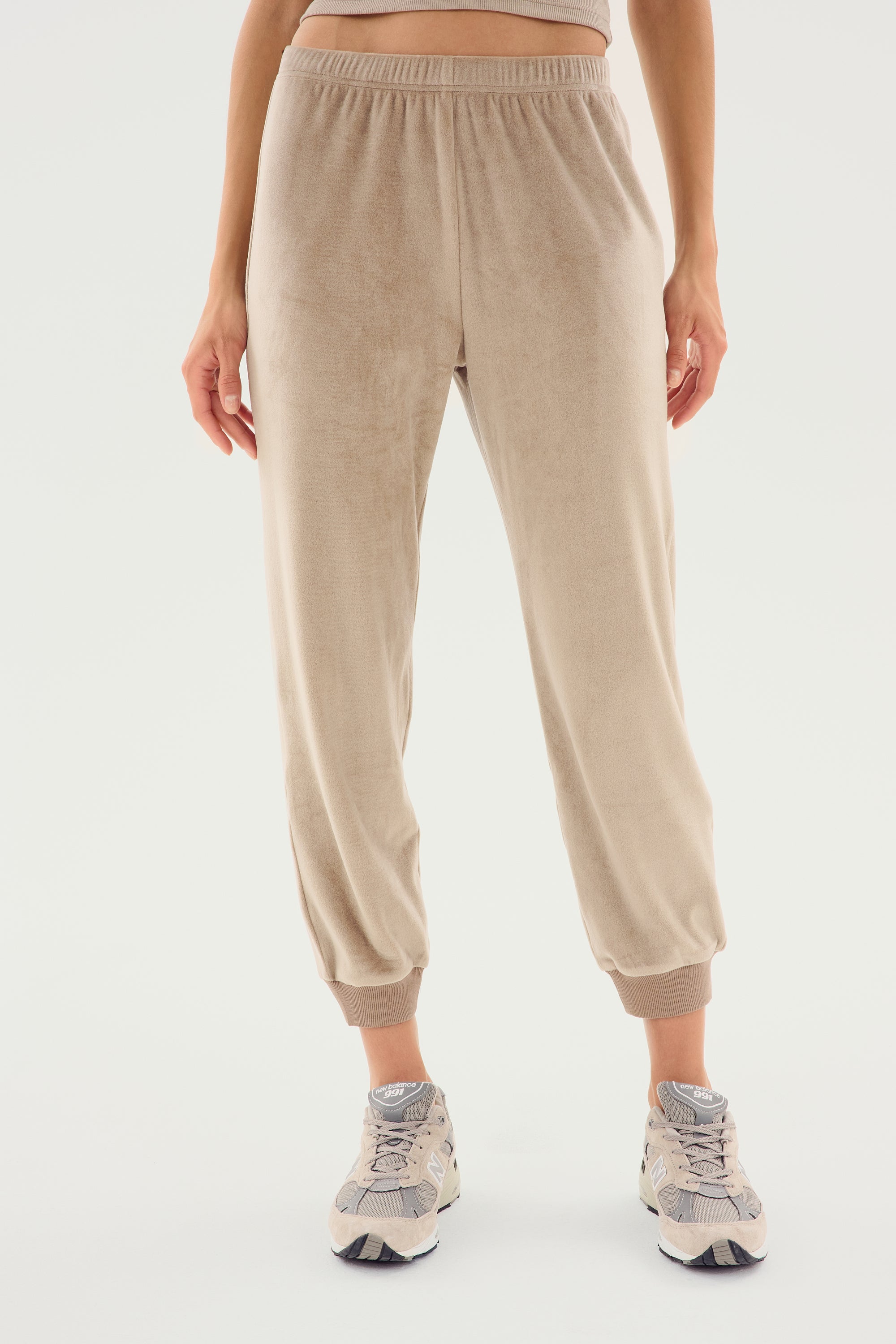 A person wearing beige Rhys Velour Jogger - Latte pants from SPLITS59 and grey sneakers stands against a plain white background, epitomizing casual wear.