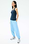 A woman stands with her hands in the pockets of light blue joggers, wearing the SPLITS59 Ashby Rib Tank in Indigo and silver sneakers against a plain white background.