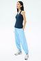 A woman stands with her hands in the pockets of light blue joggers, wearing the SPLITS59 Ashby Rib Tank in Indigo and silver sneakers against a plain white background.