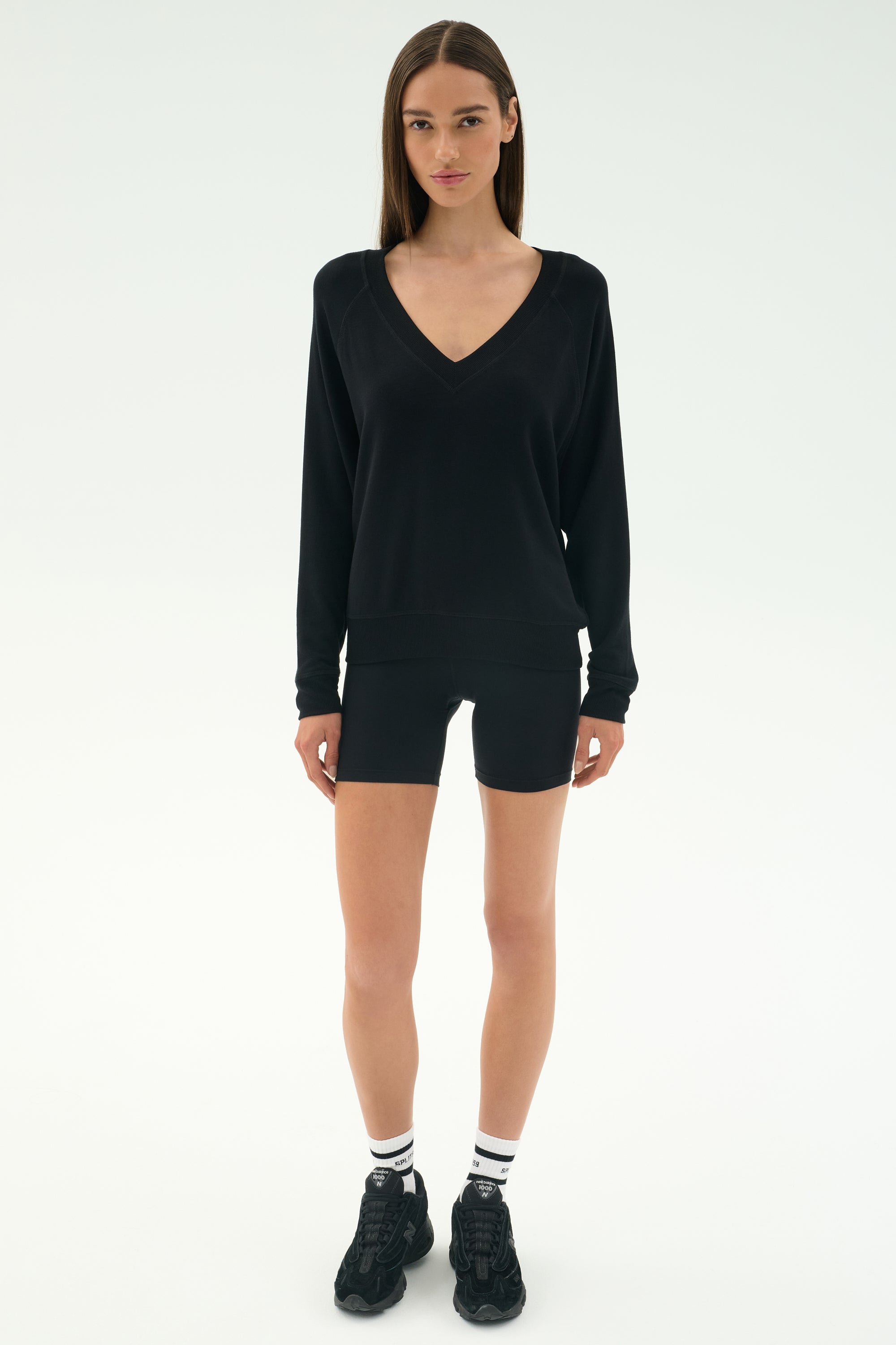 A woman faces forward against a plain light background, wearing the SPLITS59 Bennie Fleece V-Neck Sweatshirt in black, paired with black shorts, white socks, and black sneakers.