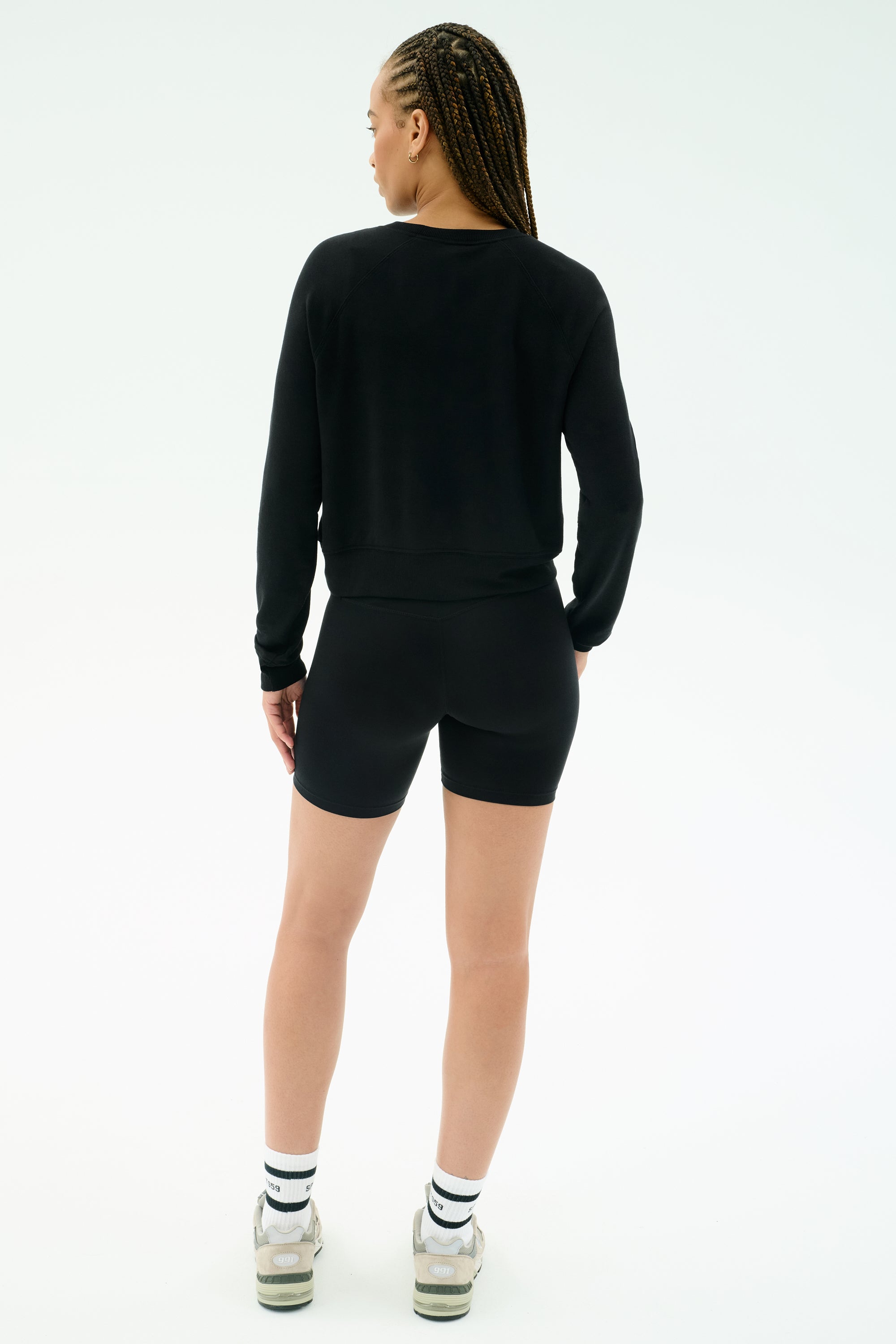 A person with braided hair stands facing away, wearing the SPLITS59 Brooks Fleece Long Sleeve Sweatshirt in black, paired with shorts, white socks, and sneakers against a plain light background. The crewneck features raglan sleeves and is made in Los Angeles.