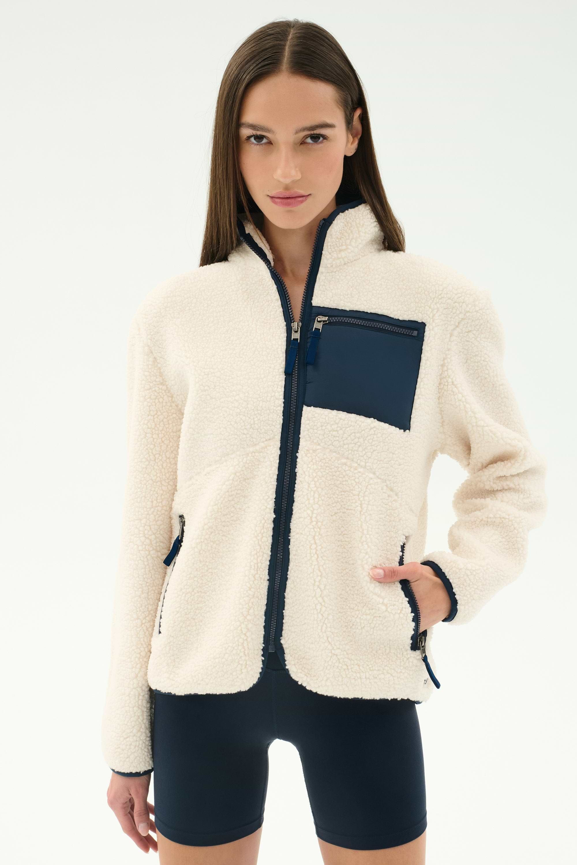 A woman models the SPLITS59 Harris Sherpa Jacket in Creme/Indigo, featuring navy zippers and front pockets, paired with black shorts against a plain white background.