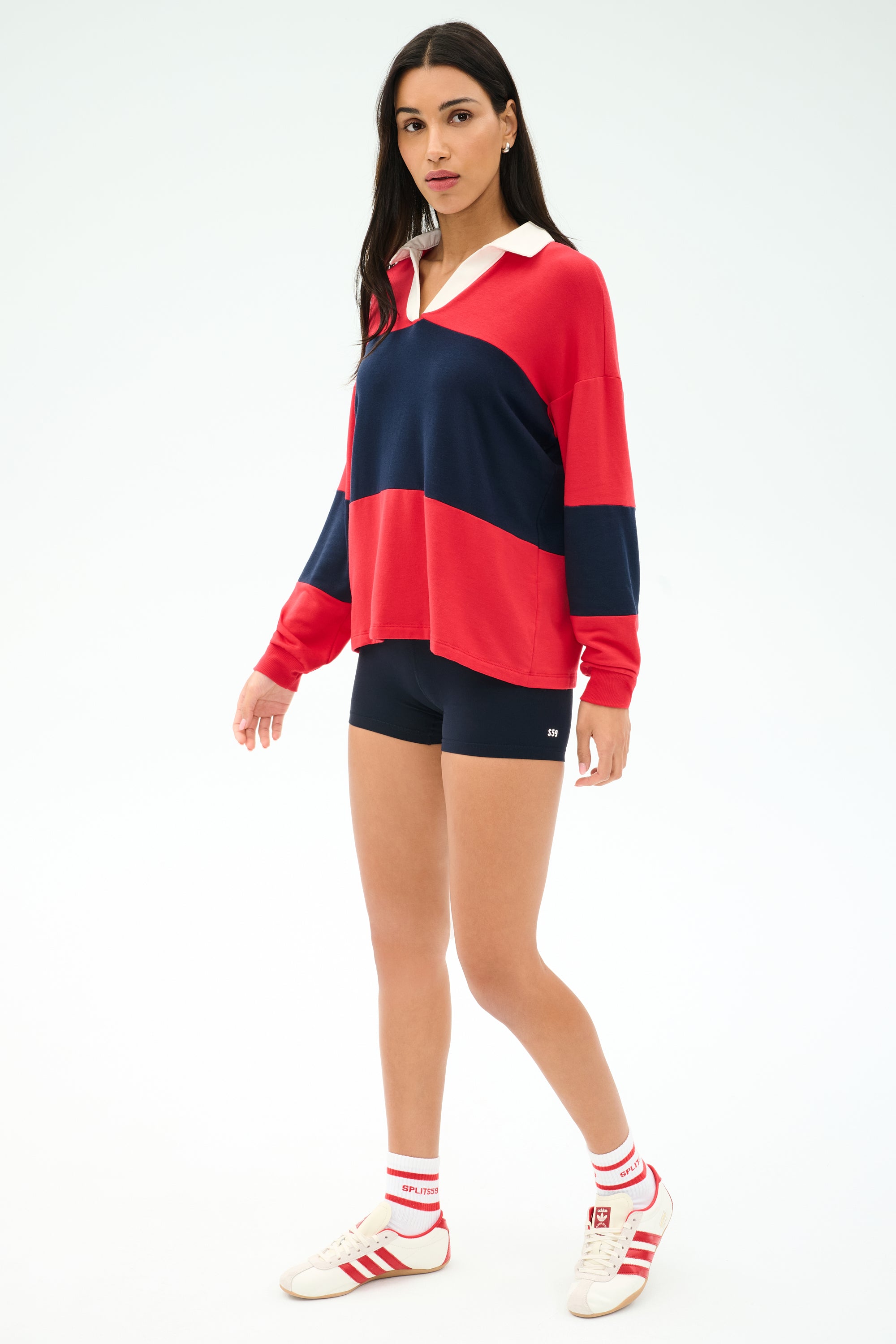 A woman stands against a white background wearing the SPLITS59 Katie Fleece Rugby in Pirate Red/Indigo, navy shorts, white socks with red stripes, and white Adidas sneakers with red accents.