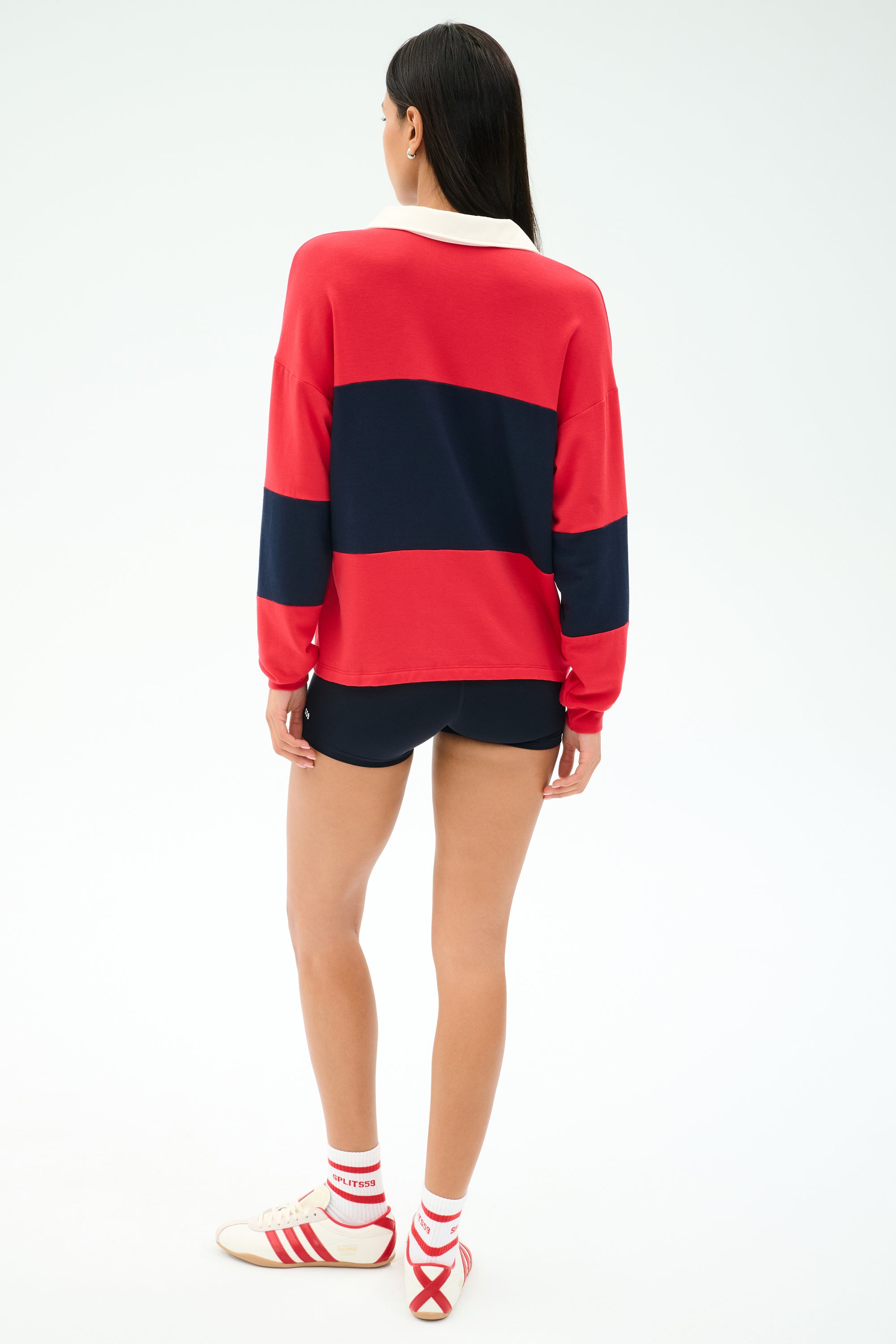 A person stands with their back to the camera, wearing the SPLITS59 Katie Fleece Rugby in Pirate Red/Indigo, black shorts, white socks, and white sneakers with red stripes.