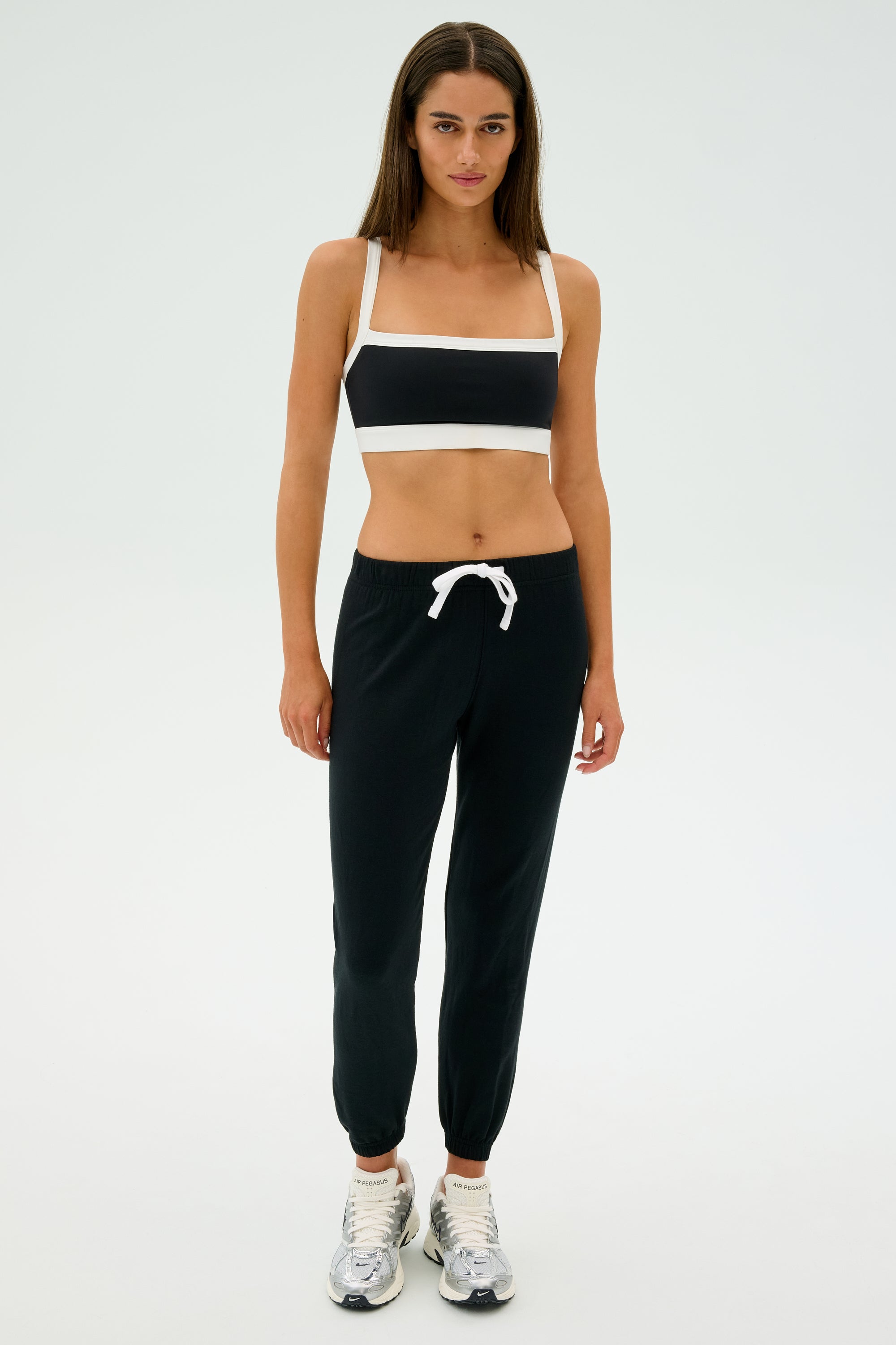 A person stands against a plain backdrop, dressed in a black and white sports bra, SPLITS59's Sonja Fleece Sweatpant in Black suited for cool weather workouts, and silver sneakers.