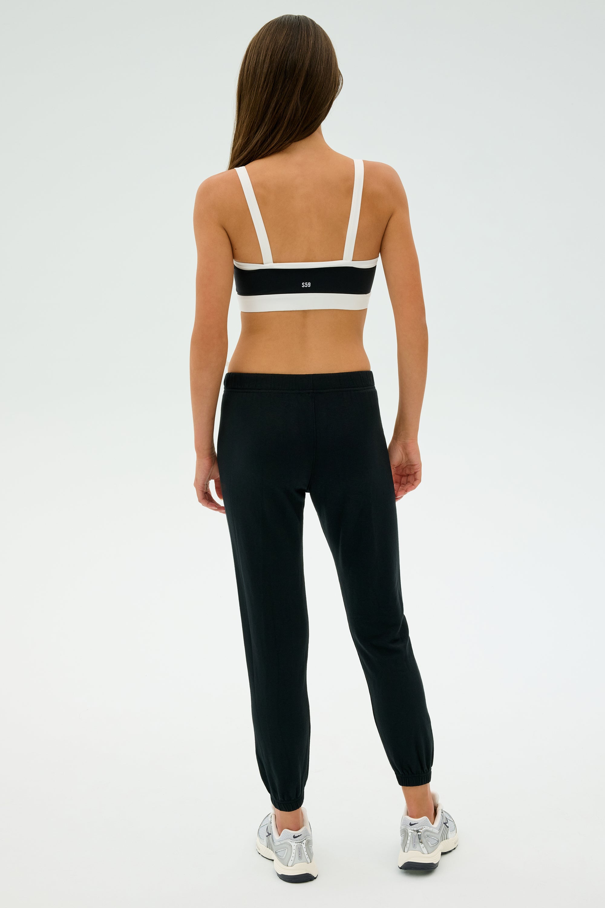 A woman facing away, wearing a black and white sports bra, Sonja Fleece Sweatpant in Black by SPLITS59, and silver sneakers, stands against a plain white background.