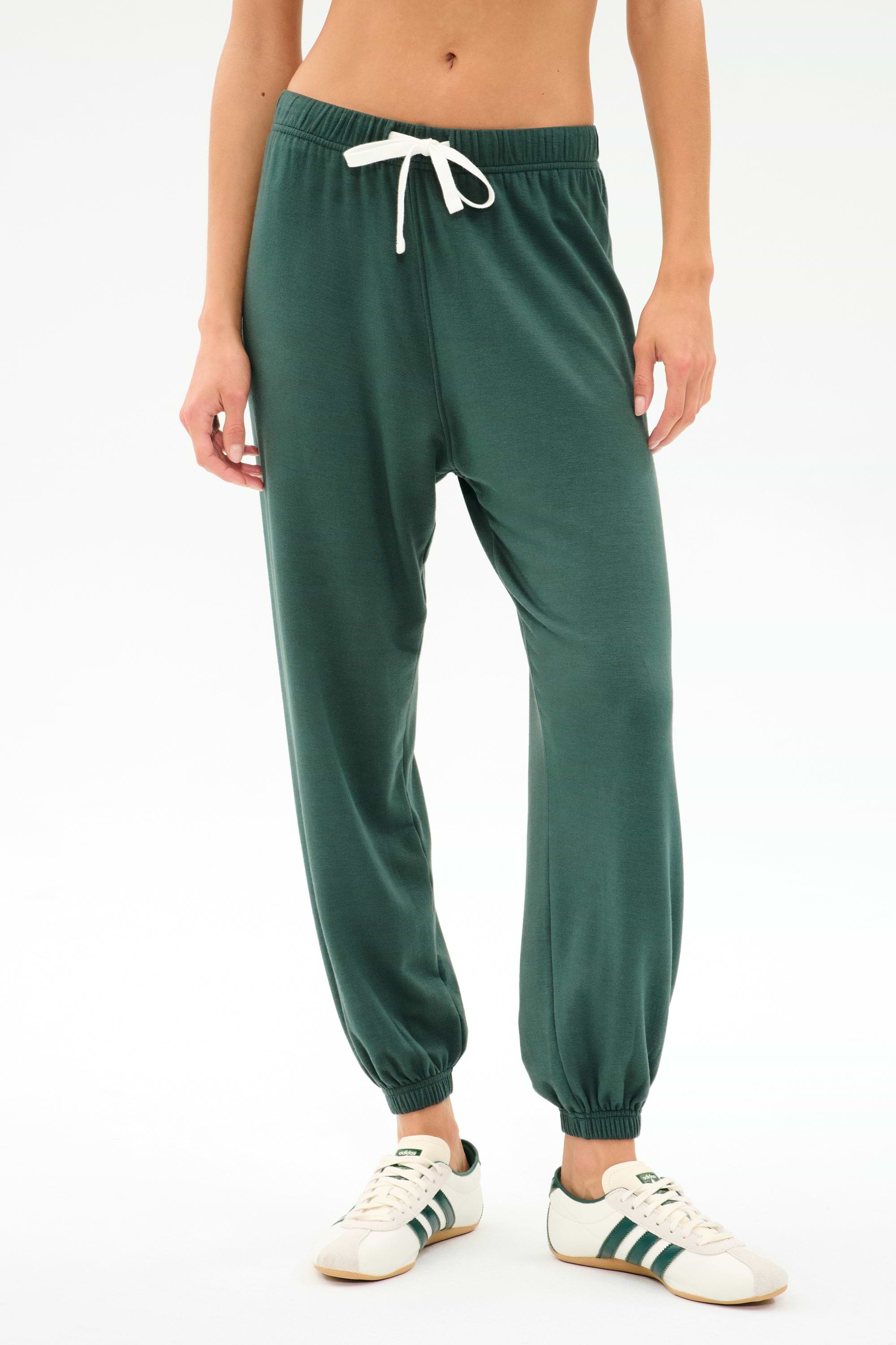 Person wears SPLITS59 Andie Oversized Fleece Sweatpant in Military, featuring a white drawstring, styled with white sneakers with green stripes—ideal for cool weather workouts—standing against a plain white background.