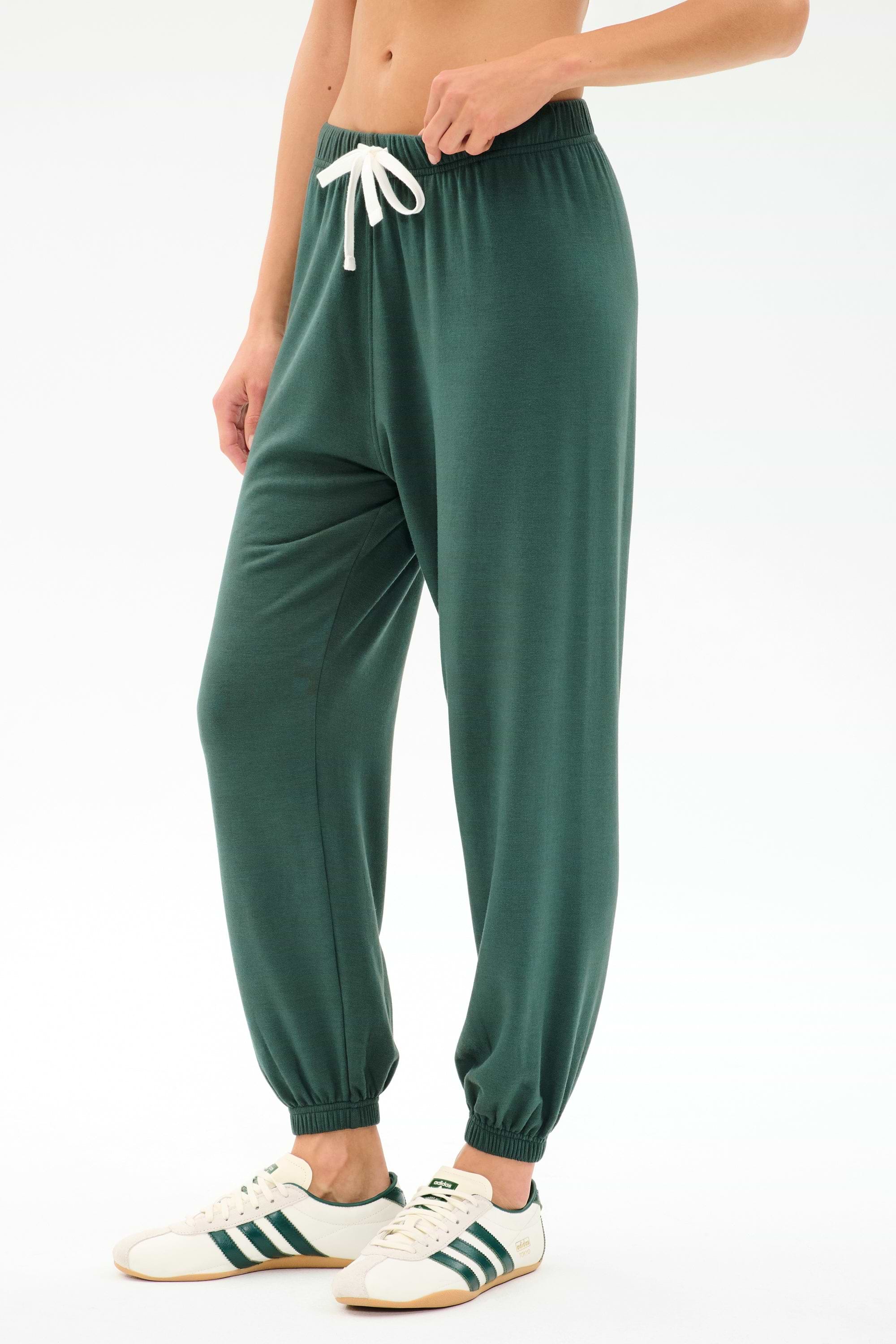 Wearing the SPLITS59 Andie Oversized Fleece Sweatpant in Military, styled with white sneakers featuring green stripes—perfect for cool weather workouts—standing against a plain white background.